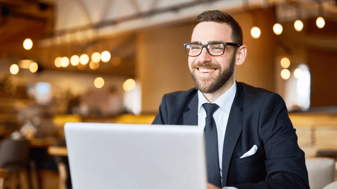 A man wearing a navy suit and tie smiles behind a laptop