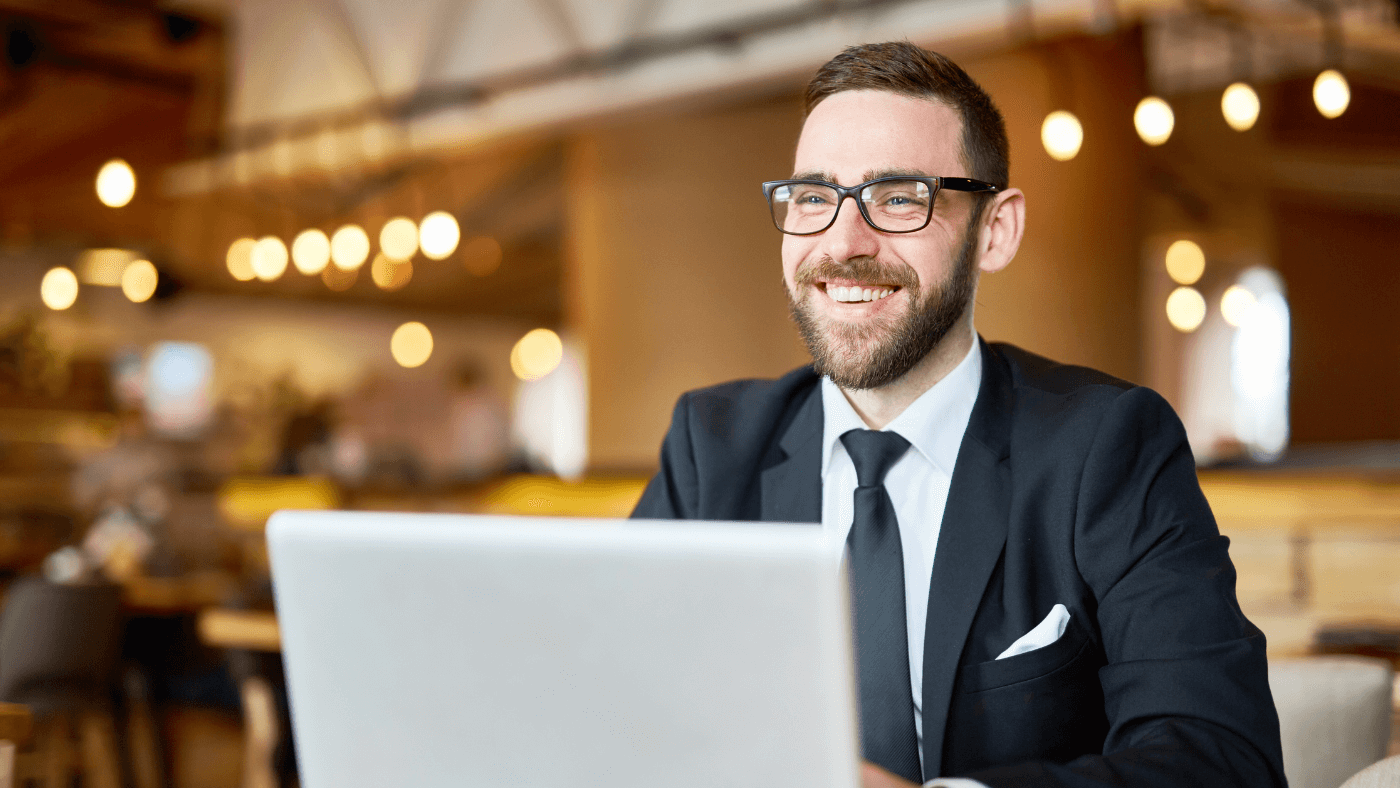 A man wearing a navy suit and tie smiles behind a laptop