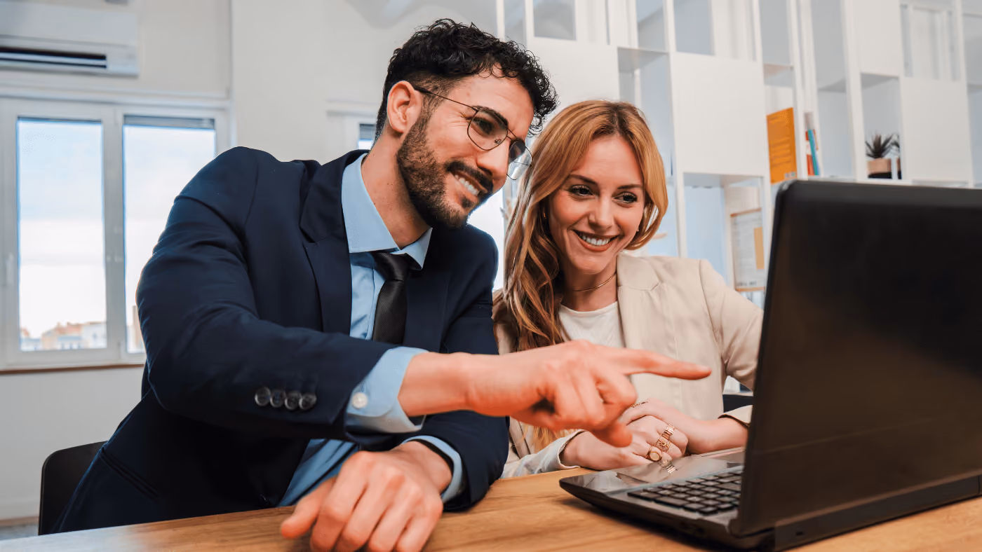 A smiling man points at a laptop while sitting next to a smiling woman in an office