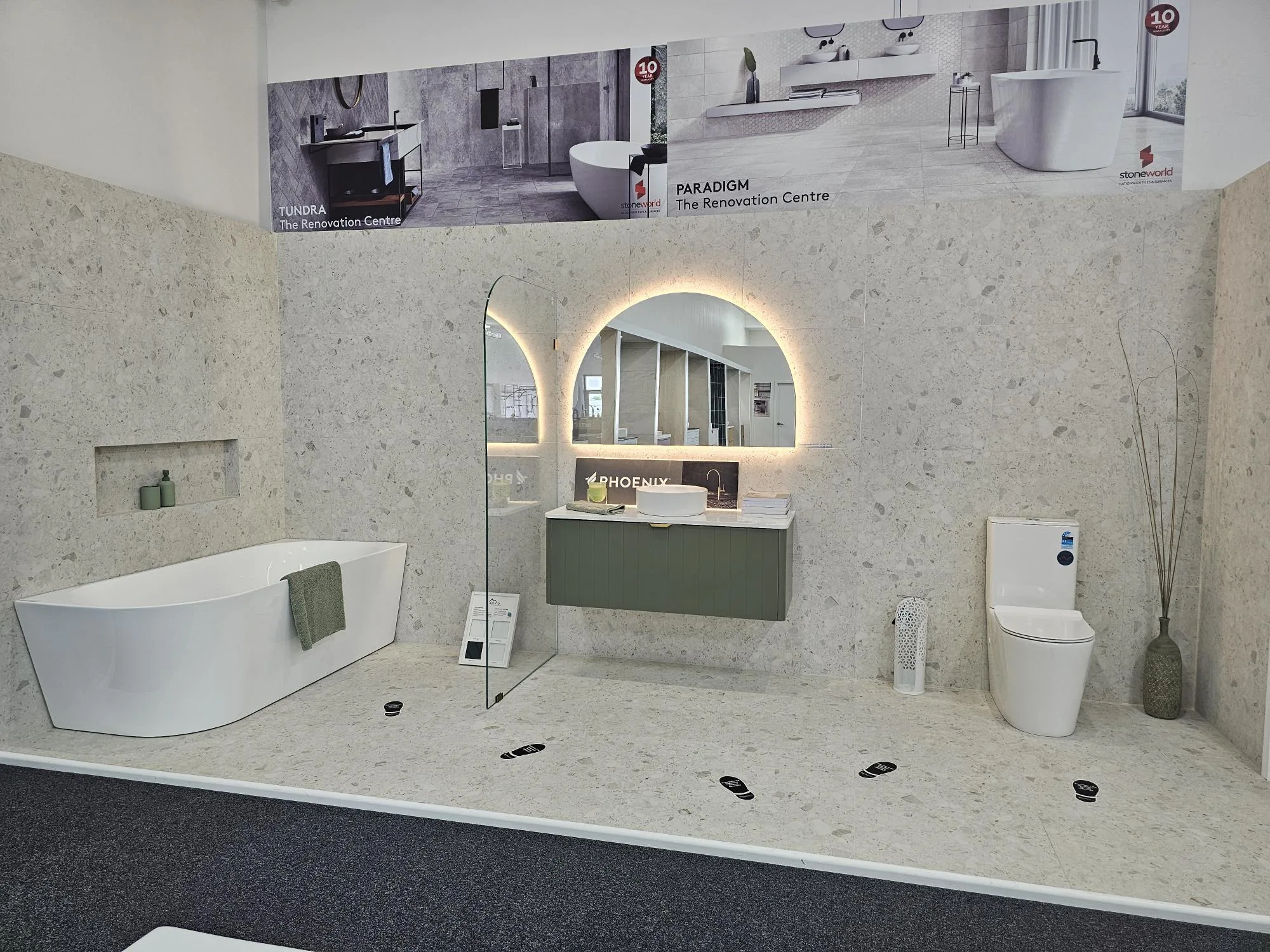Styled display bay at the Osborne Park showroom with a freestanding bath, arched backlit mirror, olive wall hung vanity and terrazzo-look wall panels