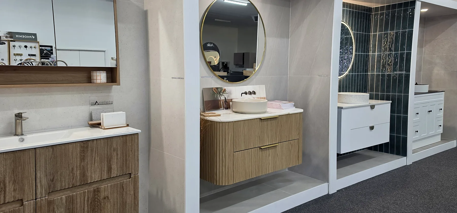 Vanity display bays in the Osborne Park showroom, a fluted timber wall hung vanity under a round brass framed mirror beside a white vanity on emerald green tiles