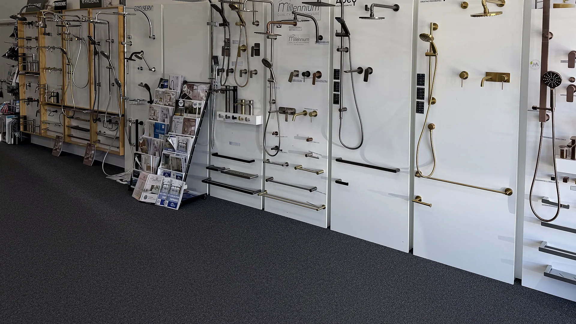 Showroom display wall of shower rails, wall mixers and heated towel rails in brushed brass, chrome and matte black under Millennium and Abey signage