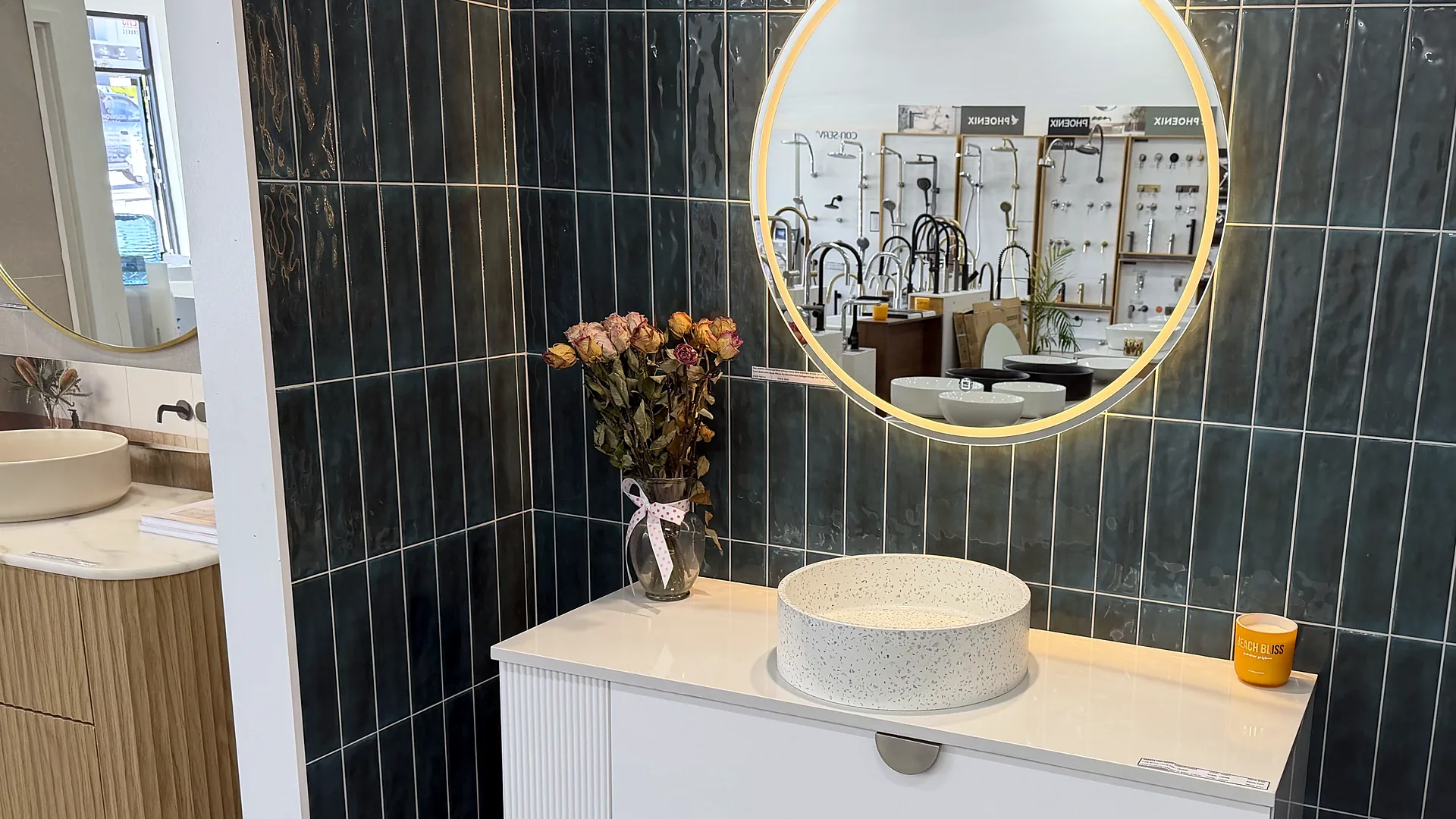 Speckled terrazzo above counter basin on a white fluted vanity against a dark green tiled wall, with a backlit round mirror and dried flowers