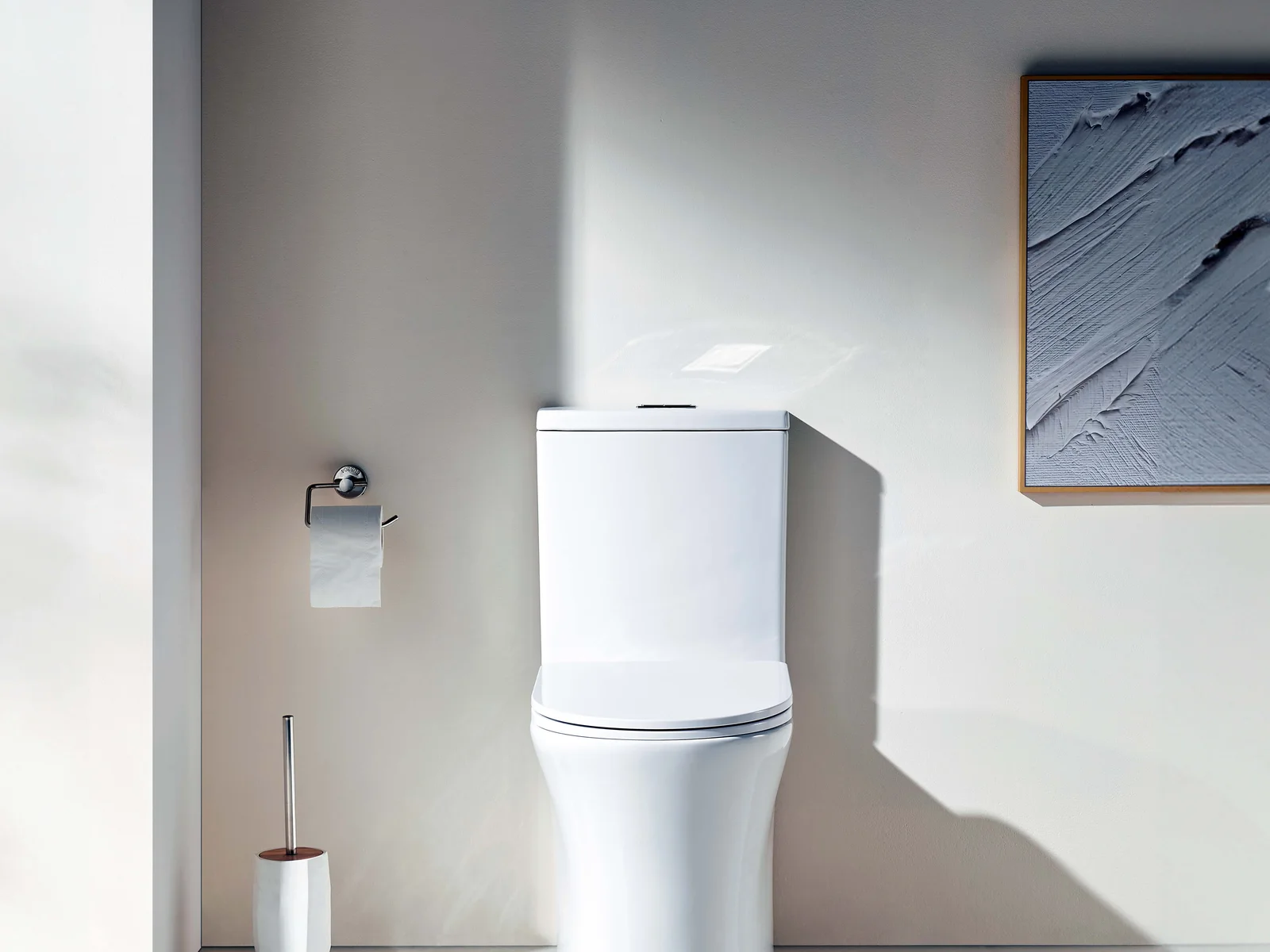 White back to wall rimless toilet suite styled in a bathroom with a chrome roll holder and a toilet brush against a warm neutral wall