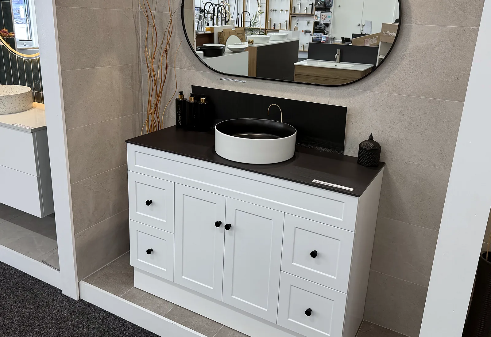 Styled display bay with a white shaker vanity, a dark stone benchtop, a matte black vessel basin and an oval mirror