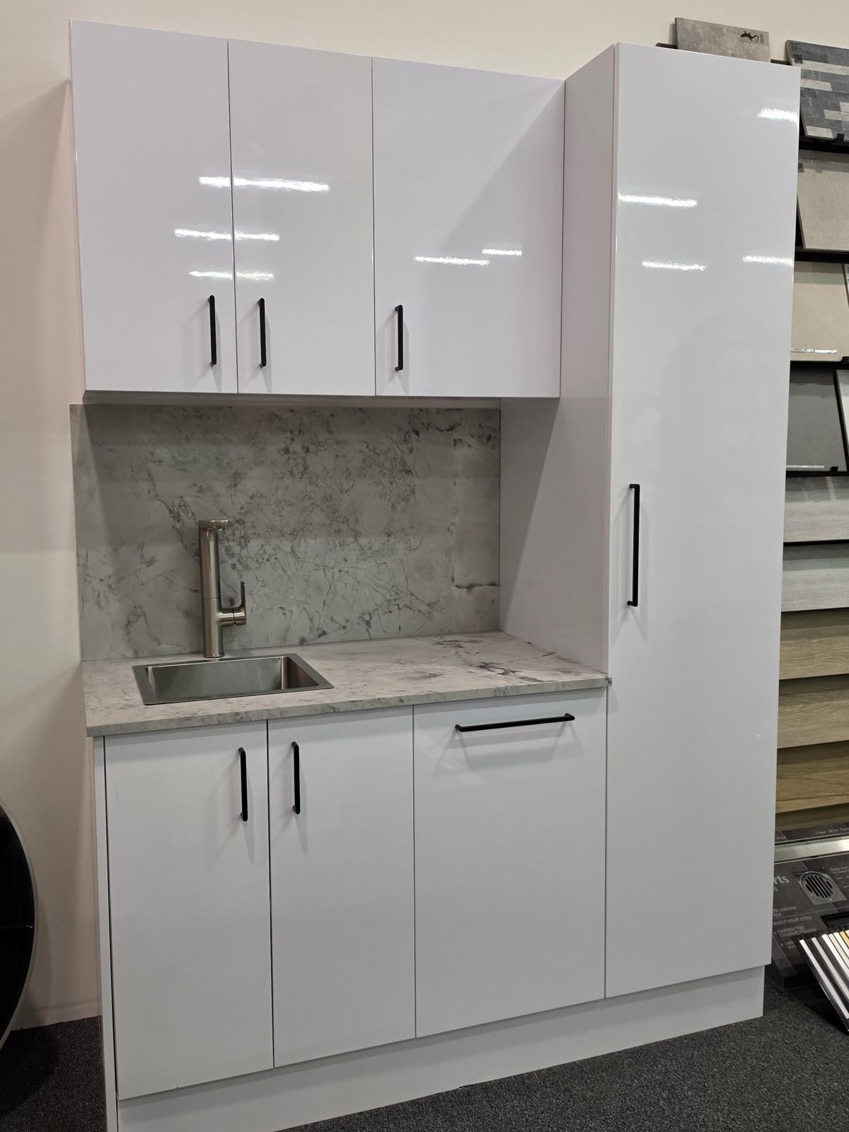 Alpine white gloss laundry unit on display in the Osborne Park showroom, with matte black bar handles, a marble look benchtop and splashback, a stainless sink and a full height broom cupboard