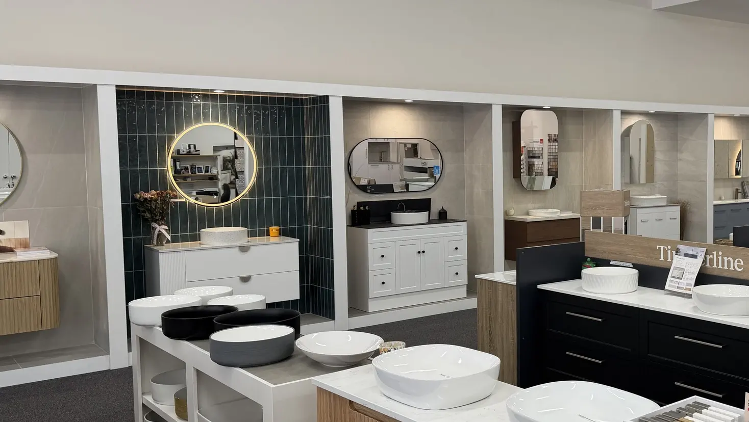 Run of vanity display bays in the Osborne Park showroom, led by a white fluted vanity with a terrazzo basin and a round backlit mirror on emerald green tiles