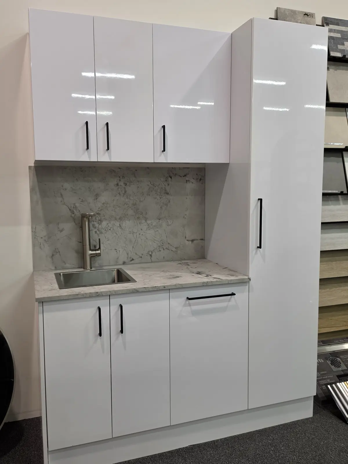 Alpine white gloss laundry unit on display in the Osborne Park showroom, with matte black bar handles, a marble look benchtop and splashback, a stainless sink and a full height broom cupboard