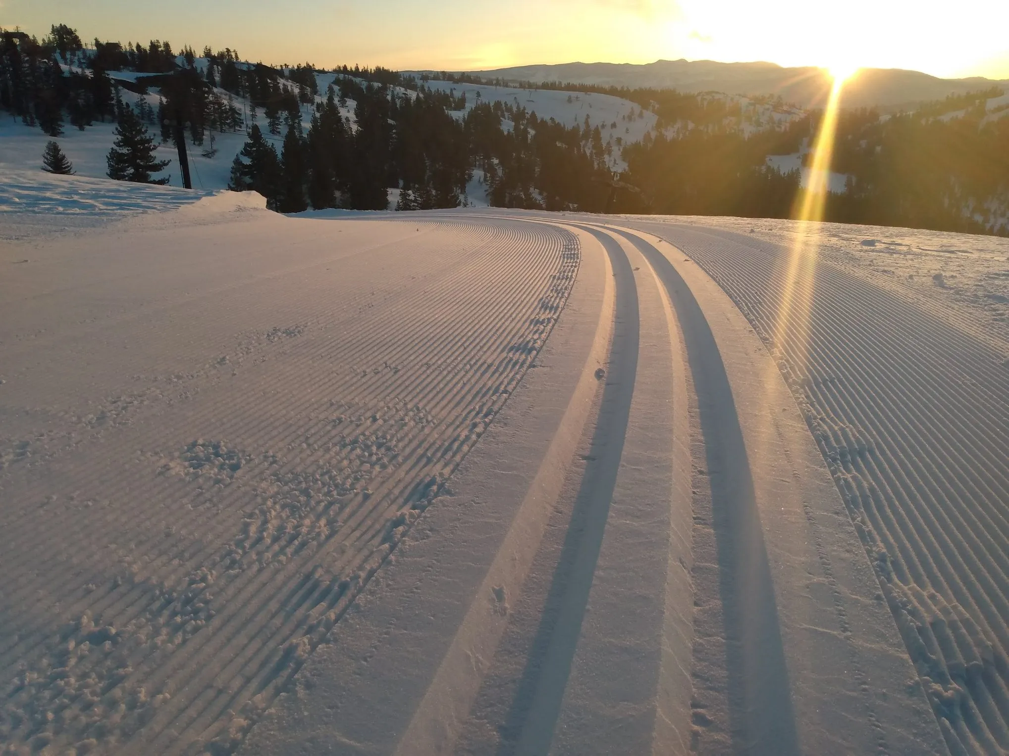 Sunset over groomed snowy cross-country ski trails with forested hills in the distance.