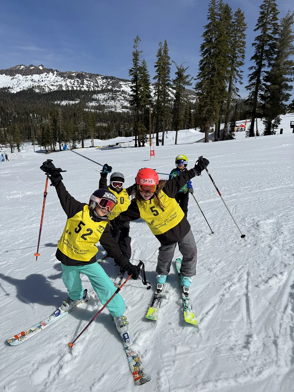 Four children wearing helmets and yellow vests posing on skis on a snowy mountain slope.
