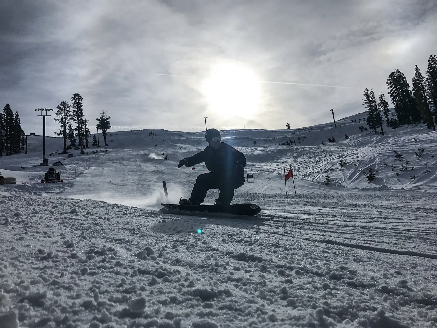 Snowboarder carving down snowy slope with sun low in cloudy sky and trees on the sides.