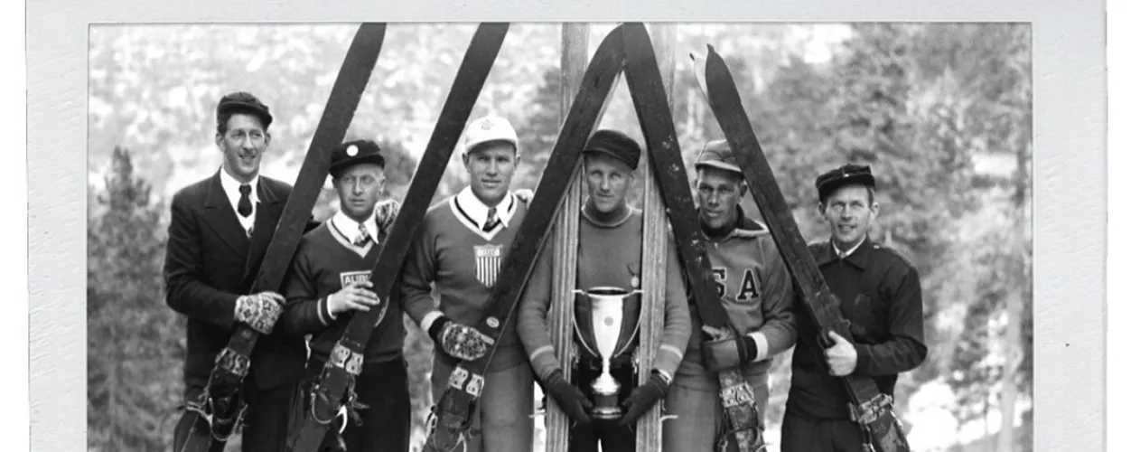 Six men in vintage skiing outfits holding skis and a trophy, posing outdoors in black and white.