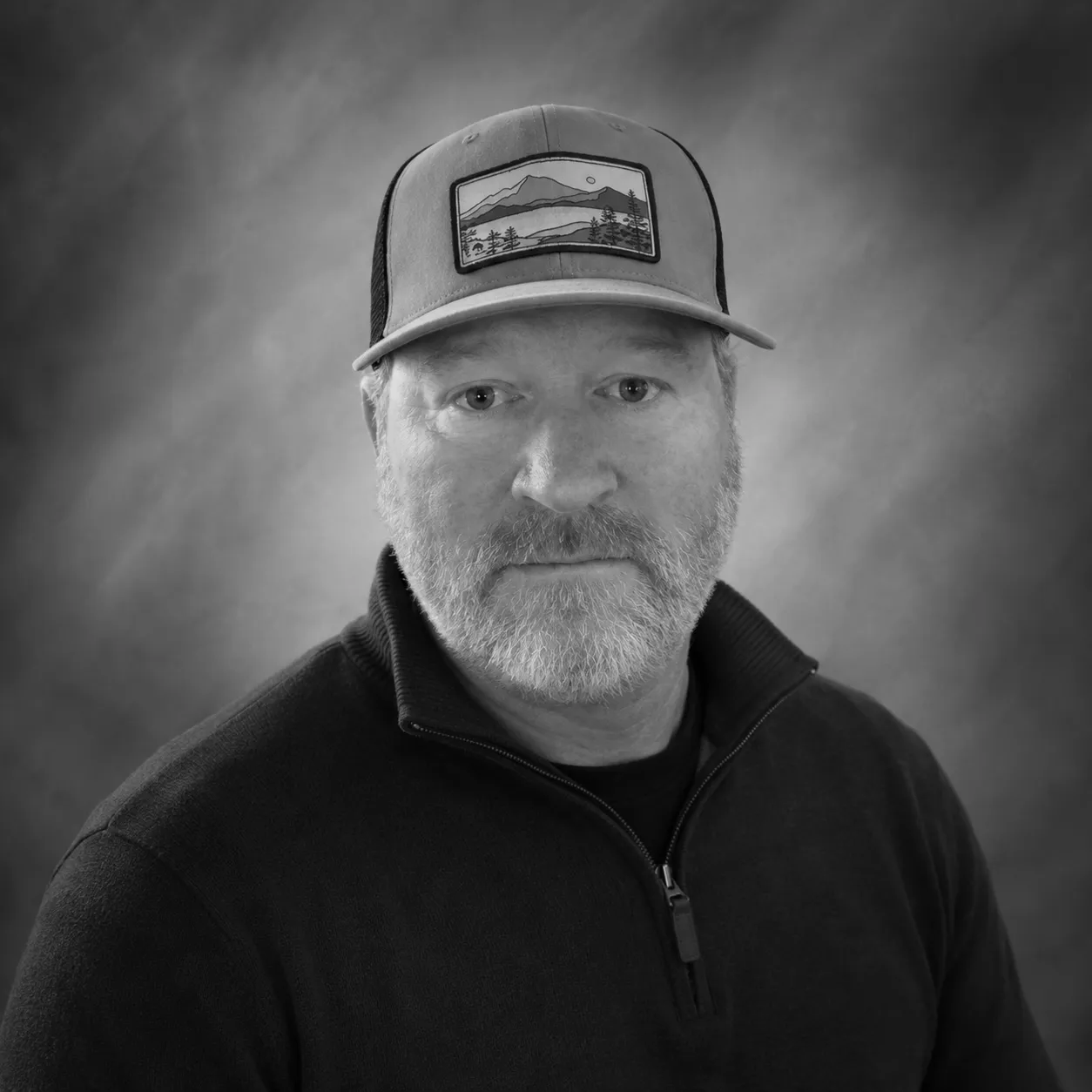 Chris Baker: Black and white portrait of a middle-aged man with a beard wearing a hat and zip-up sweater.
