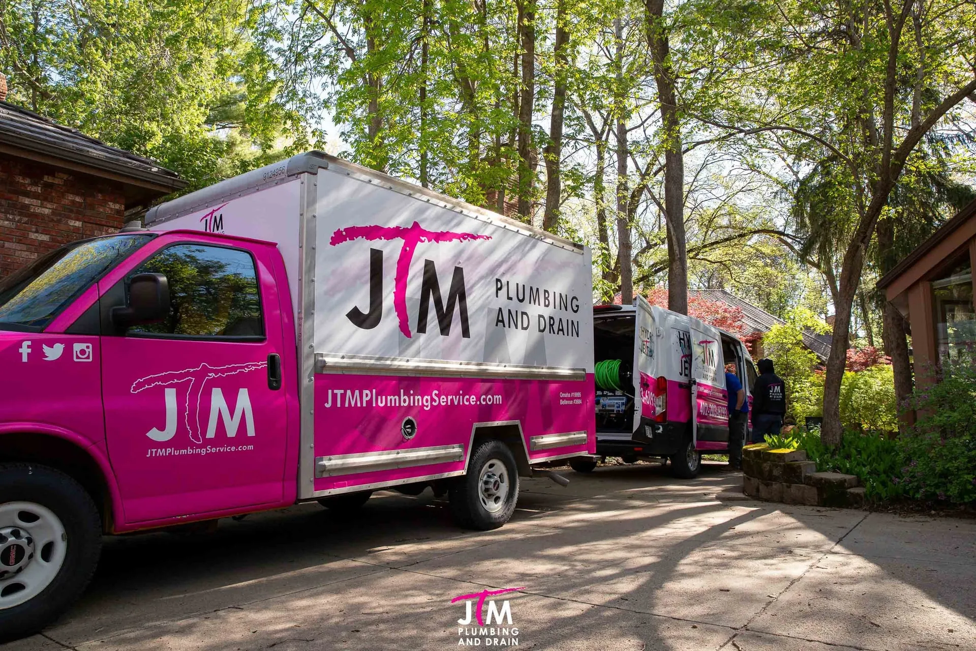 JTM Plumbing and Drain box truck and hydro jetting van parked in a wooded driveway outside a home