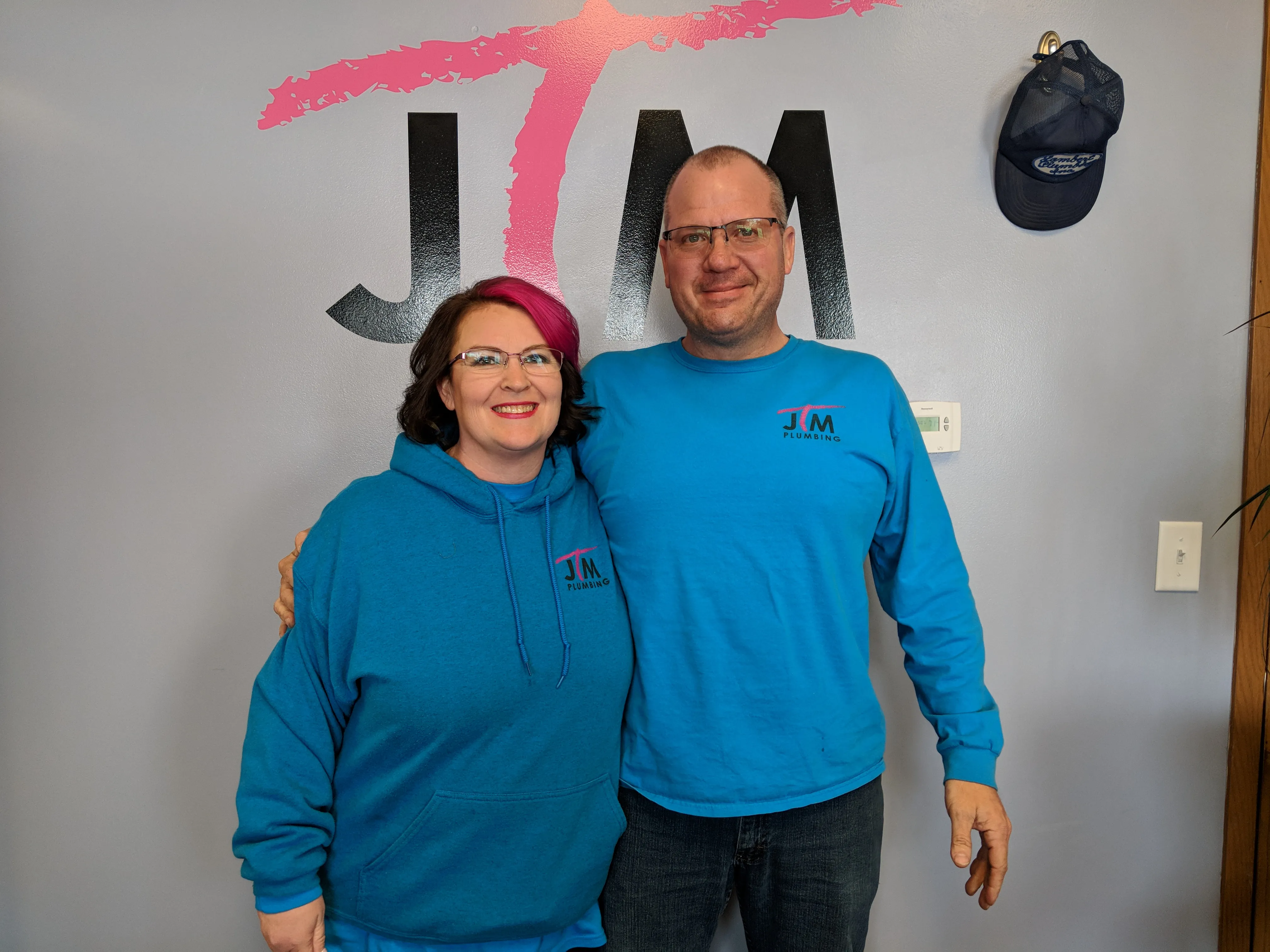 Jon and Traci Miller, owners of JTM Plumbing & Drain, standing together in front of the company logo wearing branded shirts.
