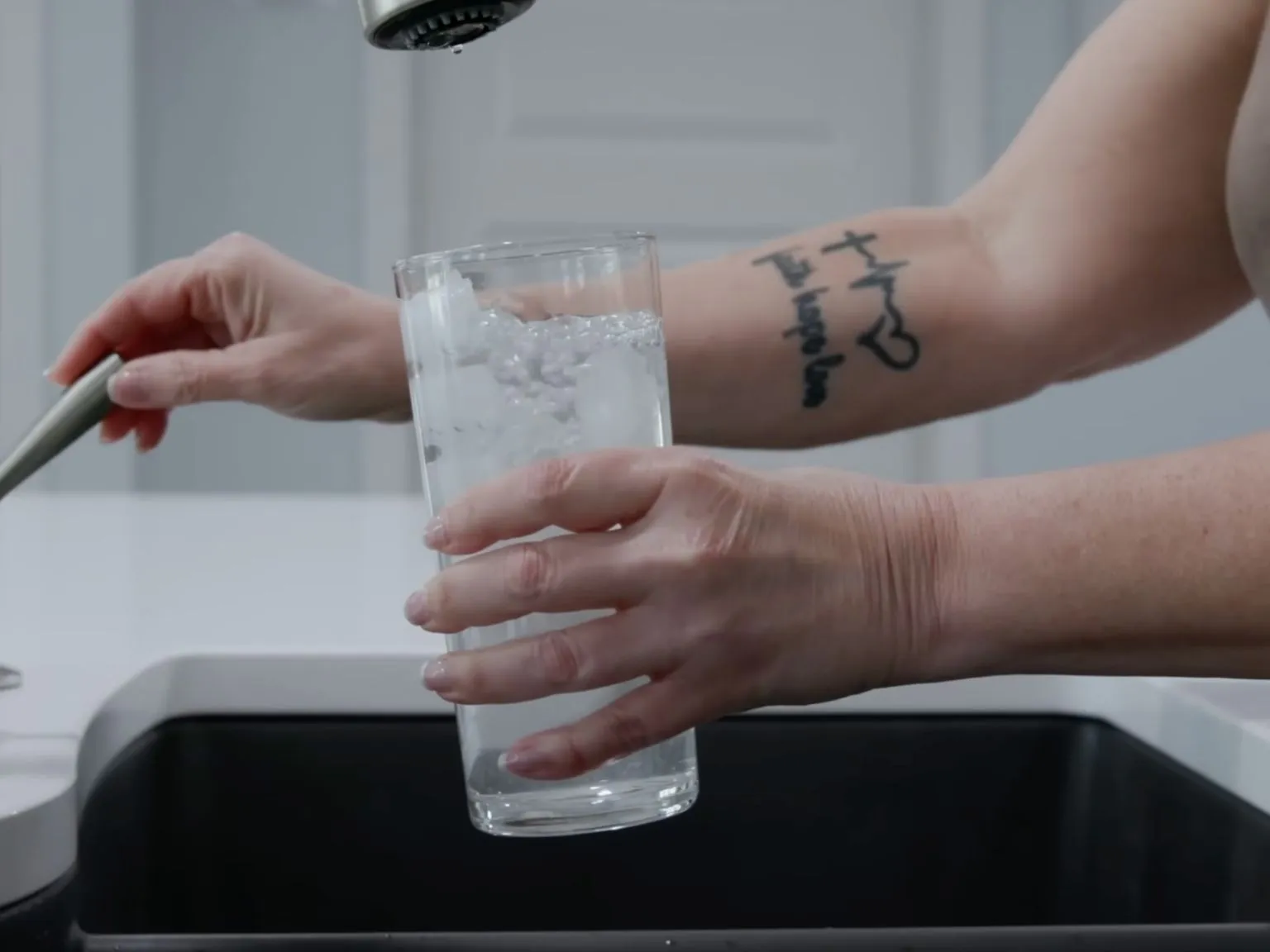 Hand filling a drinking glass with clear water from a kitchen sink faucet.