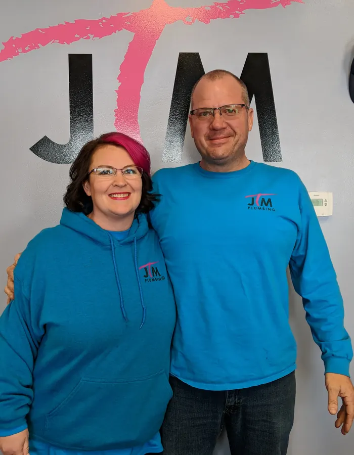 Jon and Traci Miller, owners of JTM Plumbing & Drain, standing together in front of the company logo wearing branded shirts.