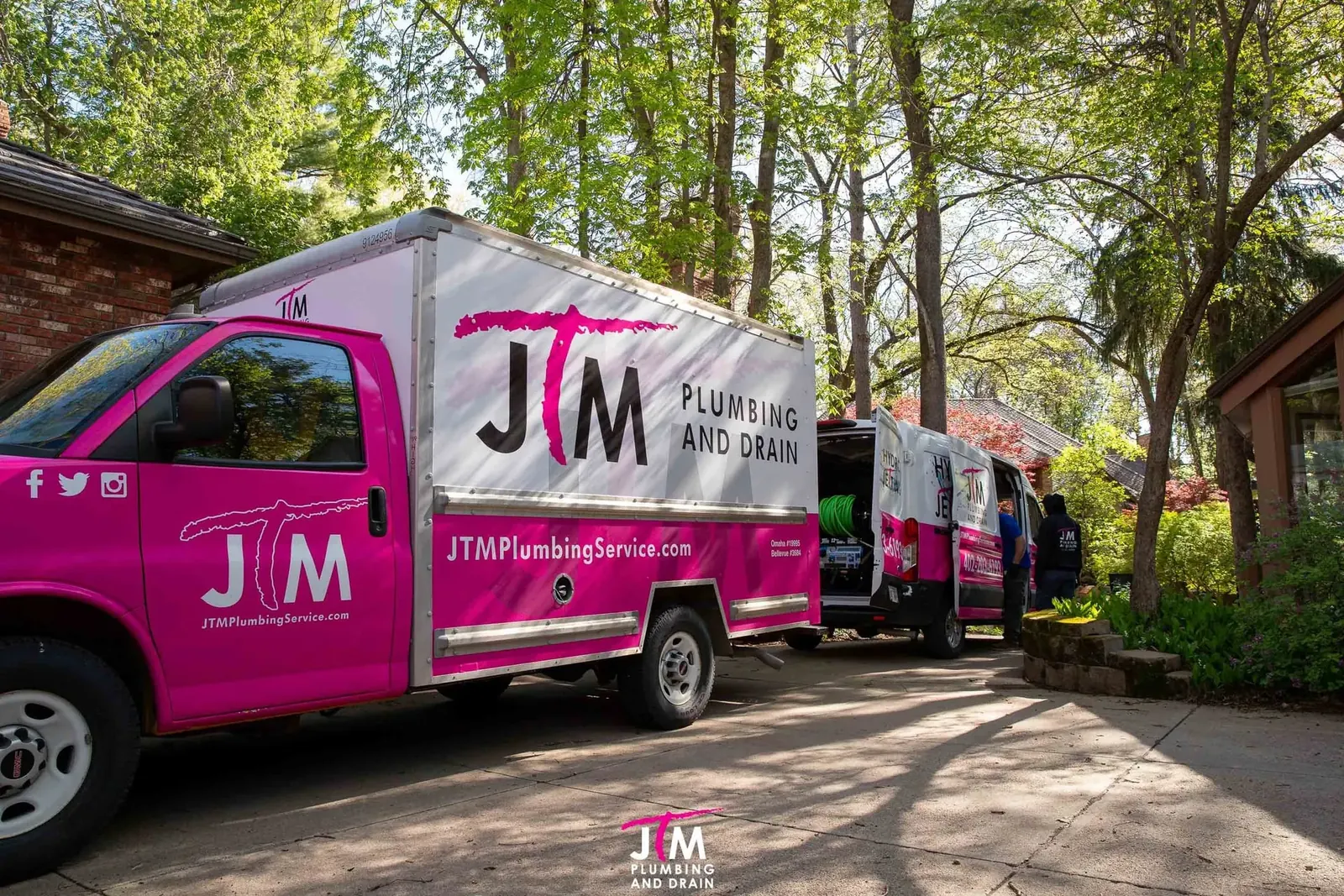 JTM Plumbing and Drain box truck and hydro jetting van parked in a wooded driveway outside a home.