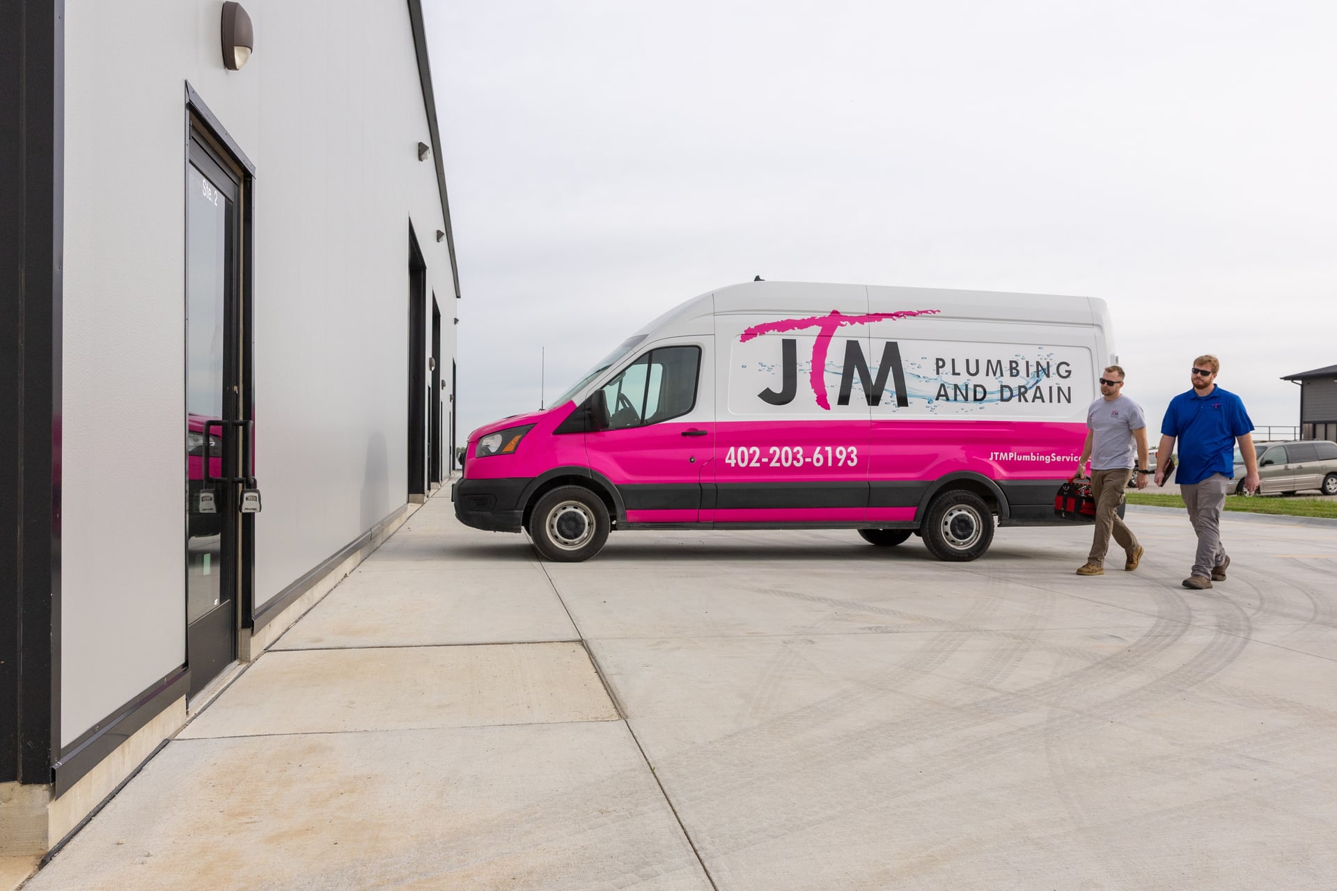 Two JTM Plumbing & Drain technicians walking toward their pink and white service van outside a building.