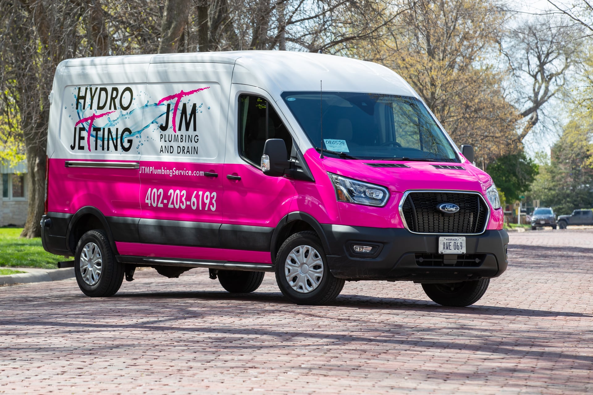 Pink and white JTM Plumbing & Drain hydro jetting van parked on a paver driveway lined with mature trees.