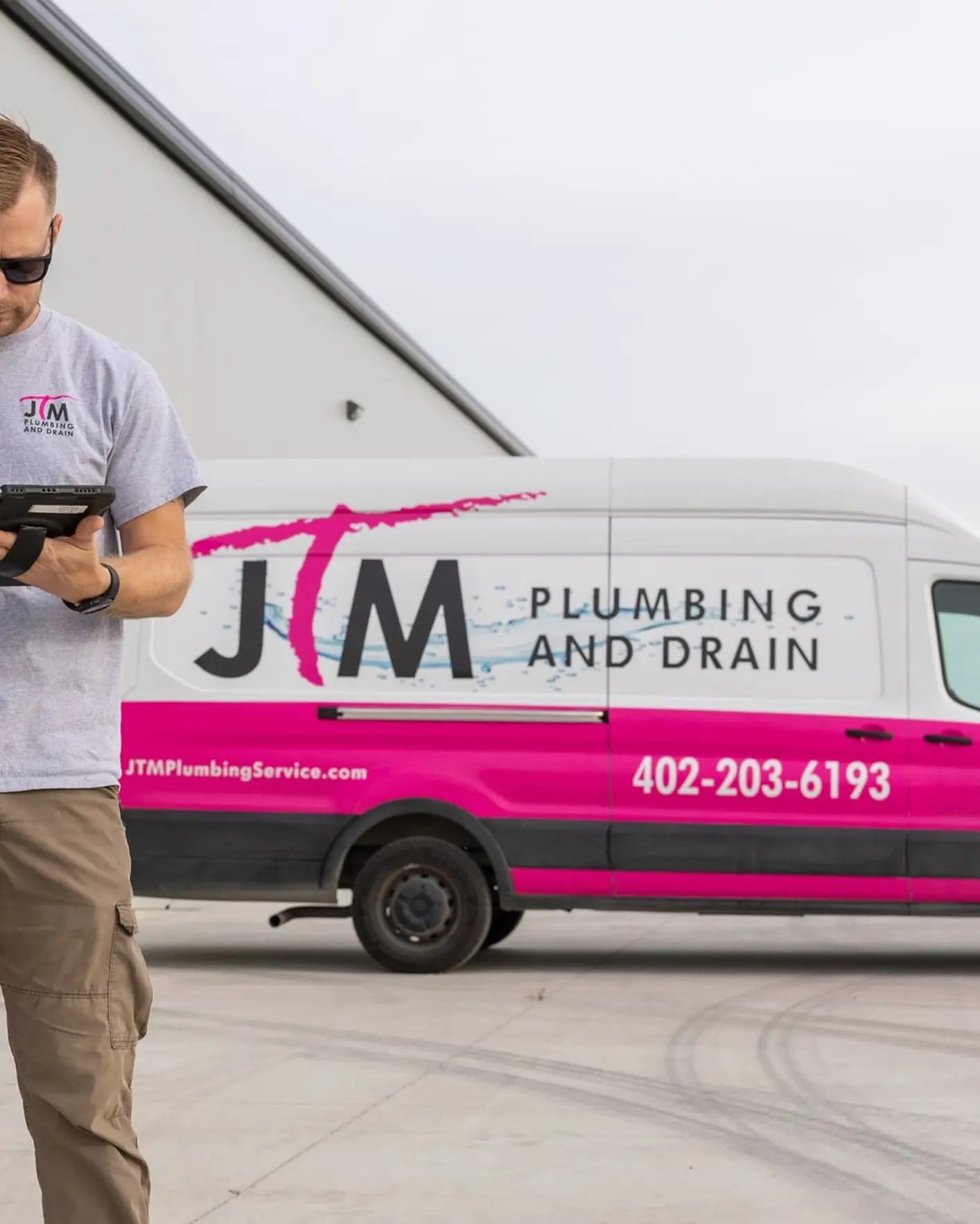 JTM Plumbing & Drain technician using a tablet outside the branded service van near a warehouse.
