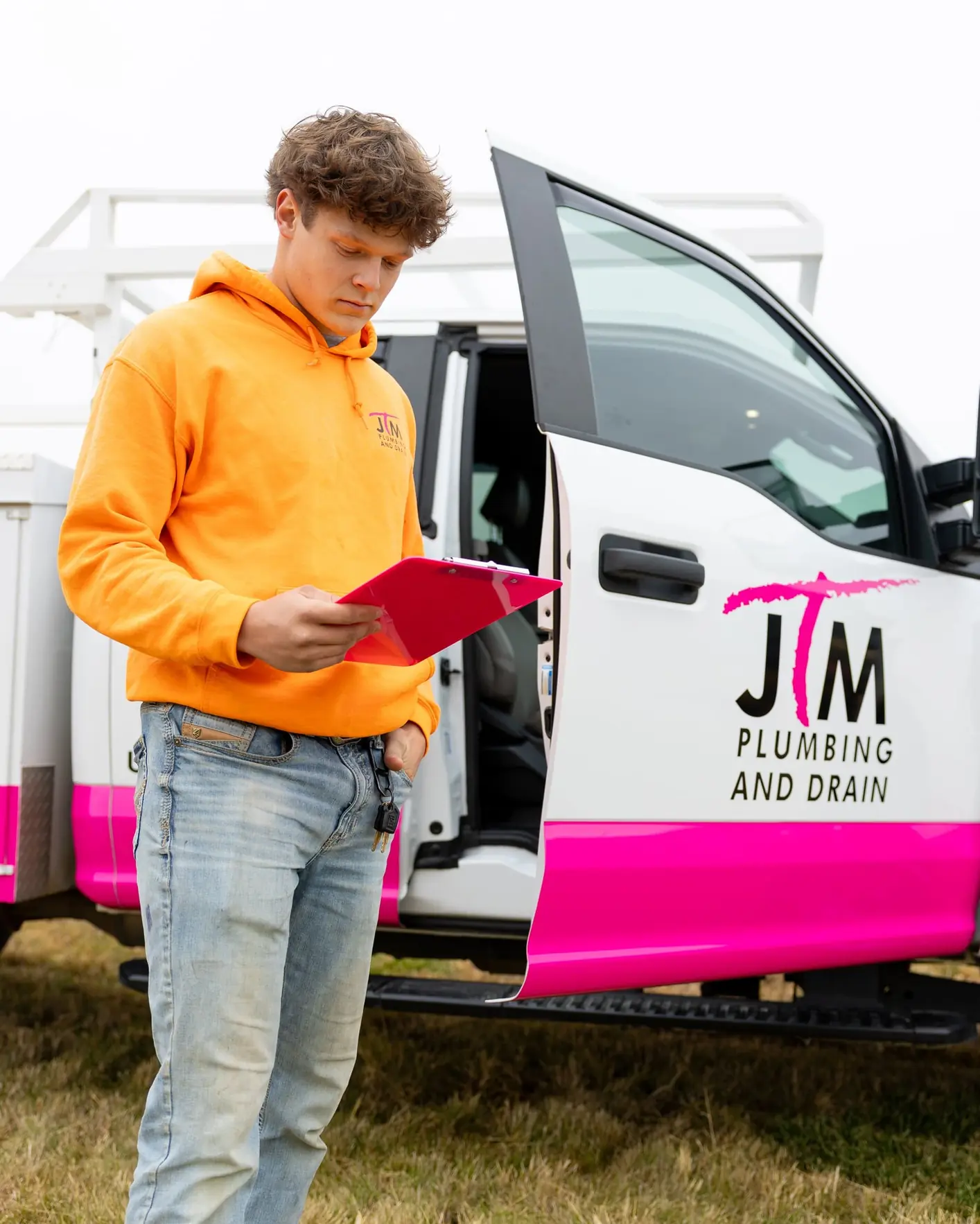 JTM Plumbing & Drain technician in an orange hoodie reviewing a work order beside the open service truck door.