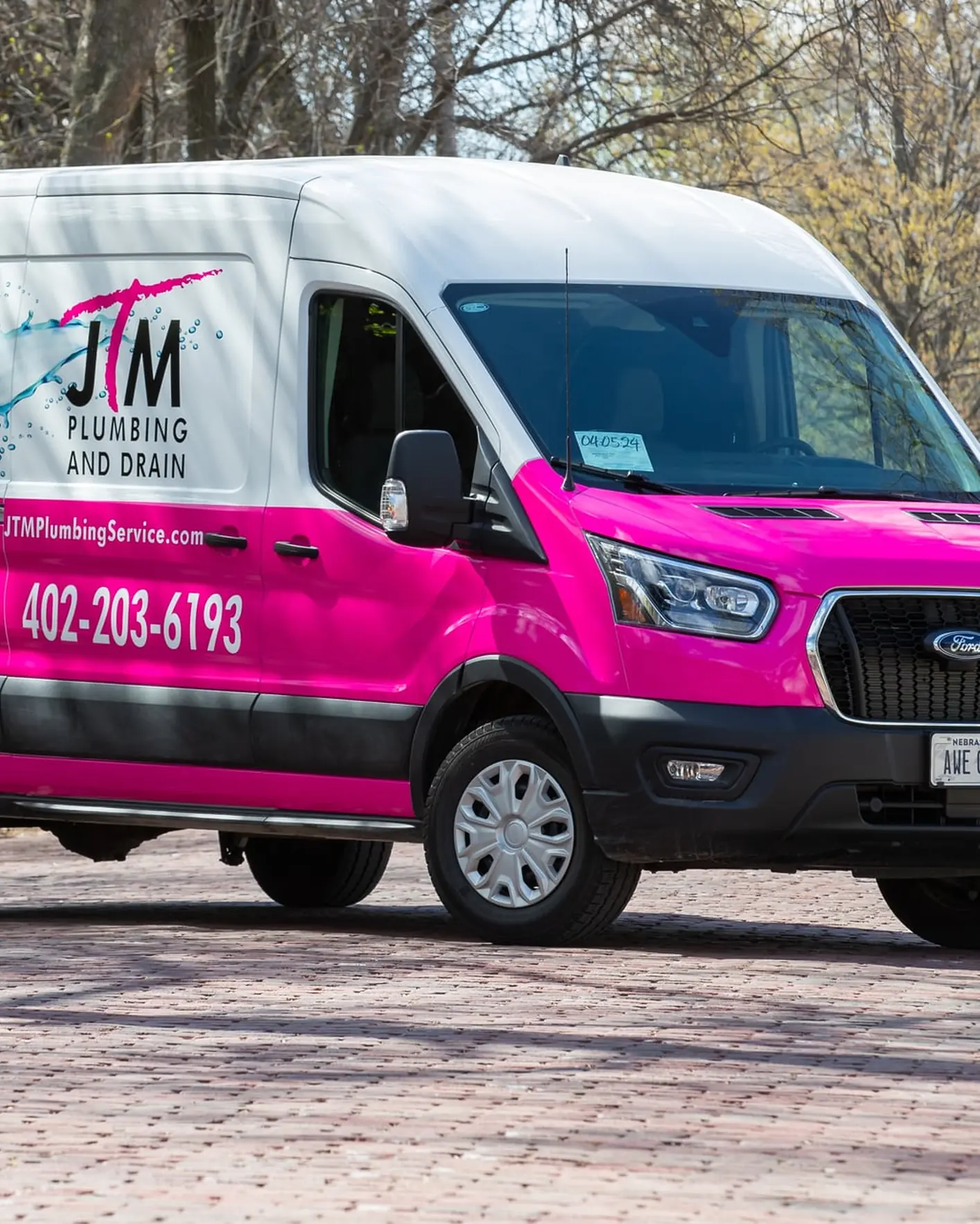 Pink and white JTM Plumbing & Drain van parked on a brick driveway in a leafy neighborhood.