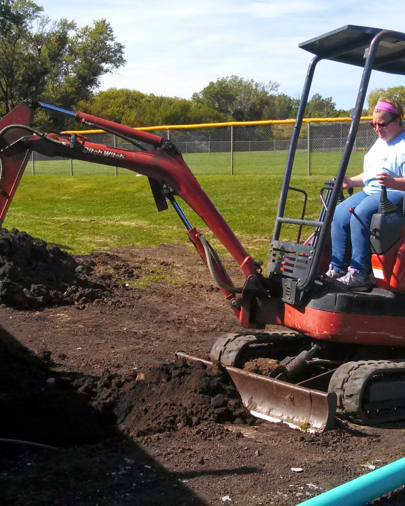 A mini excavator digs a trench beside a ball field, with a blue drain pipe laid nearby.