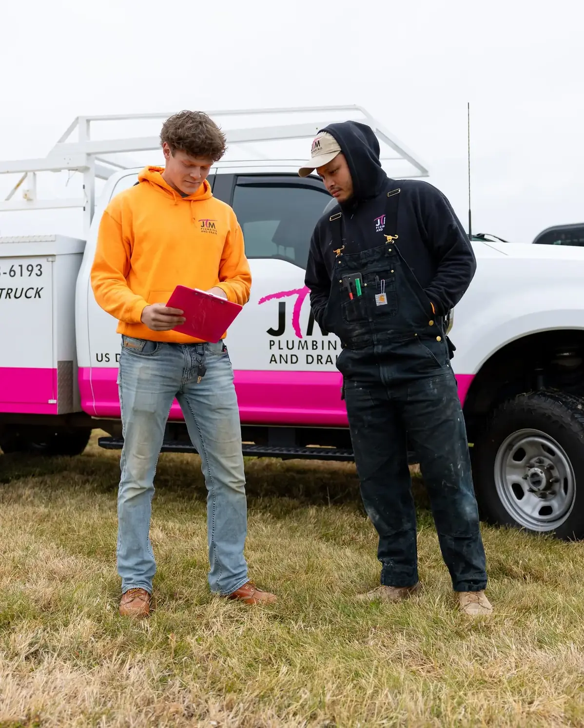 Two JTM Plumbing & Drain technicians review job notes on a clipboard beside the company truck.