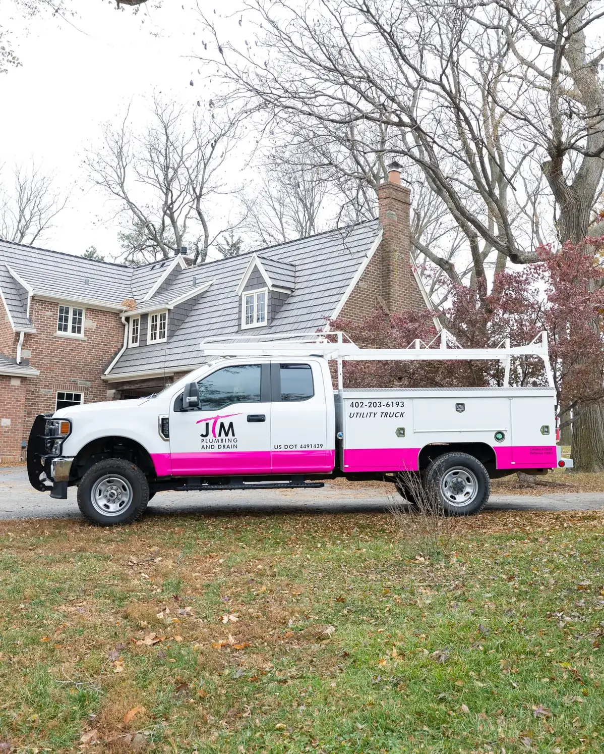 JTM Plumbing & Drain utility truck parked outside a large brick home with bare fall trees.