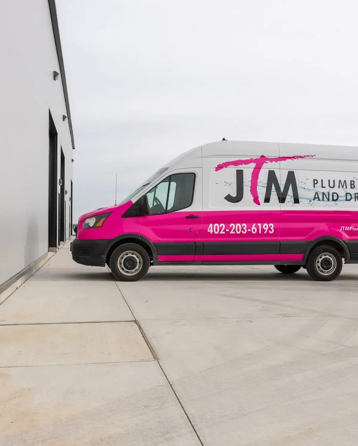 Two JTM Plumbing & Drain technicians walking outside a commercial building beside the branded pink service van.
