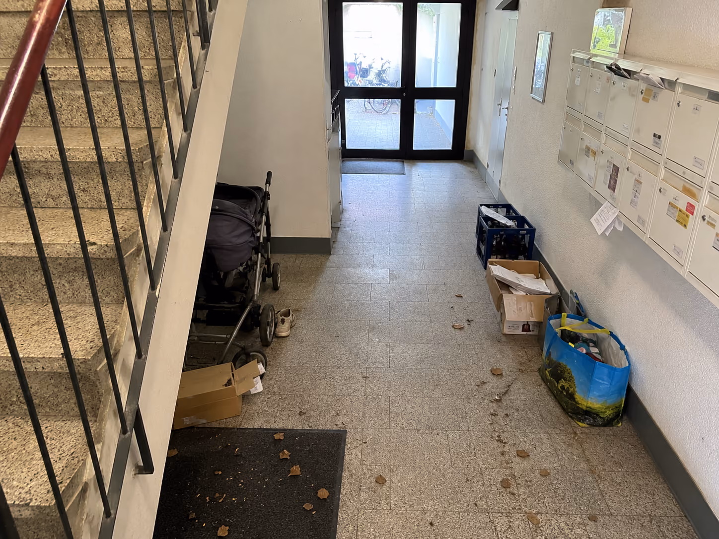 Building entry with stairs, mailboxes, a stroller, scattered leaves, and boxes on the floor.