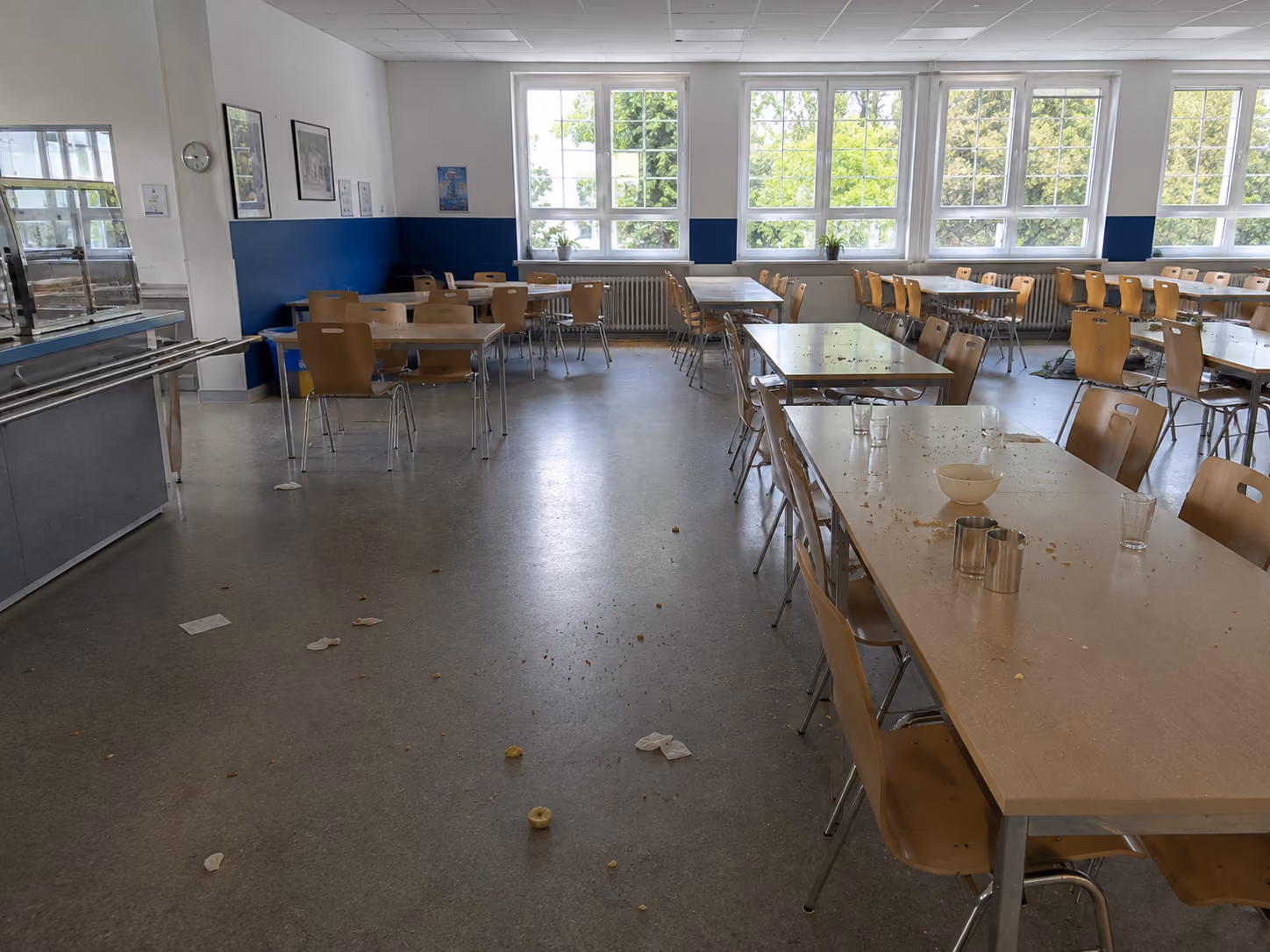 Empty school cafeteria with scattered crumbs, papers on floor, and tables with chairs under windows.