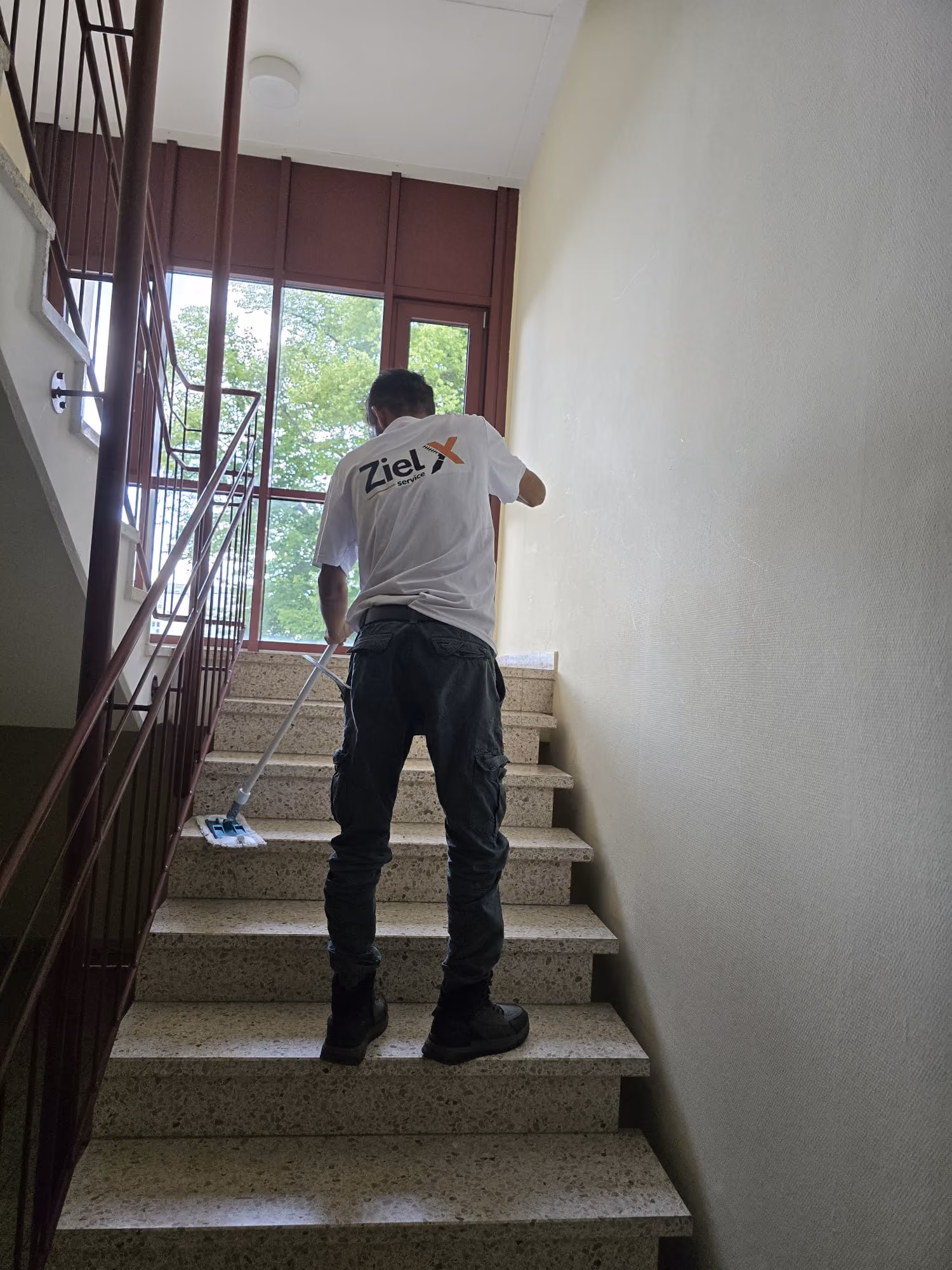 Person mopping indoor stairway with large windows and green trees outside.