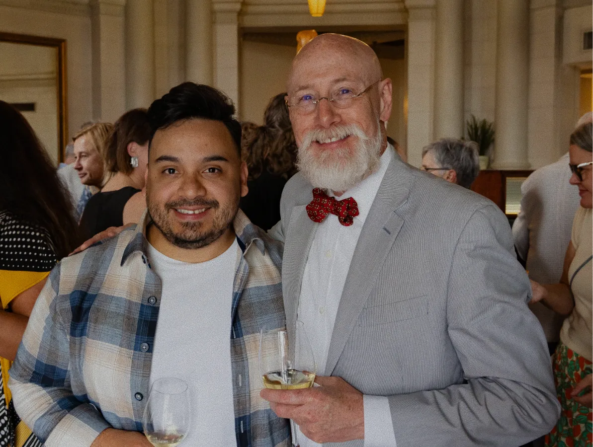 Two men smiling at a social event, one in a plaid shirt, the other in a gray suit with red bow tie.