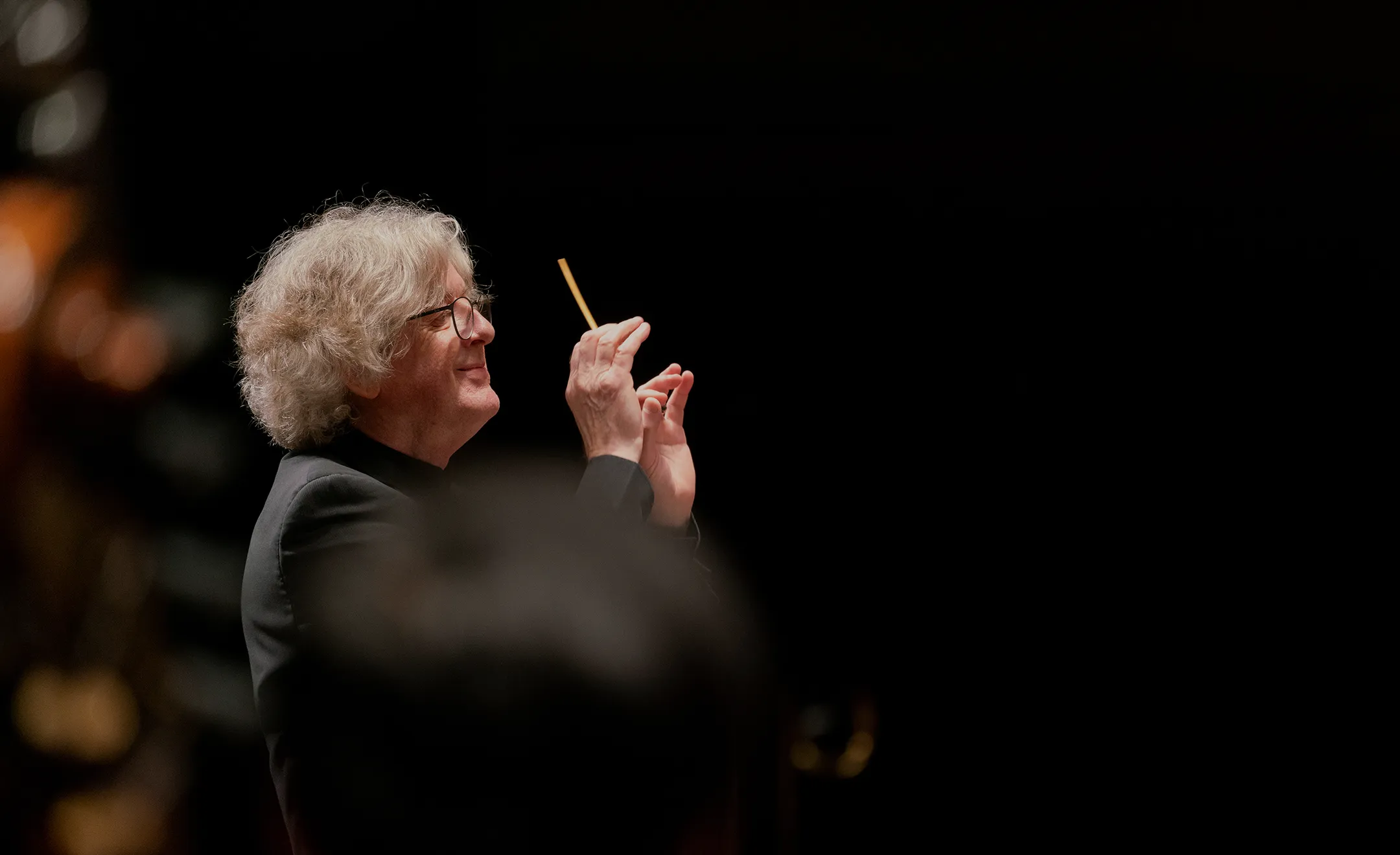 Conductor with gray curly hair and glasses holding a baton facing left, against a dark background.