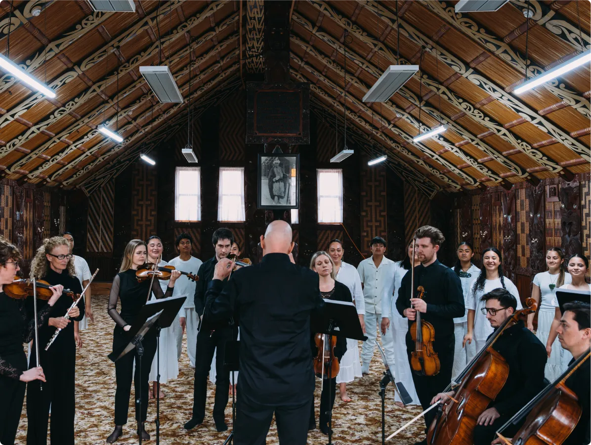 Conductor directing orchestra and choir inside a decorated hall with patterned wooden ceiling and walls.