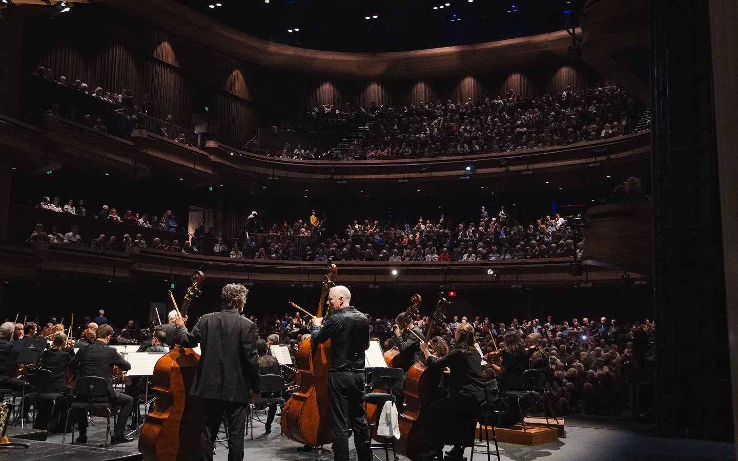 Orchestra performing on stage with an audience seated in balconies and the main floor.