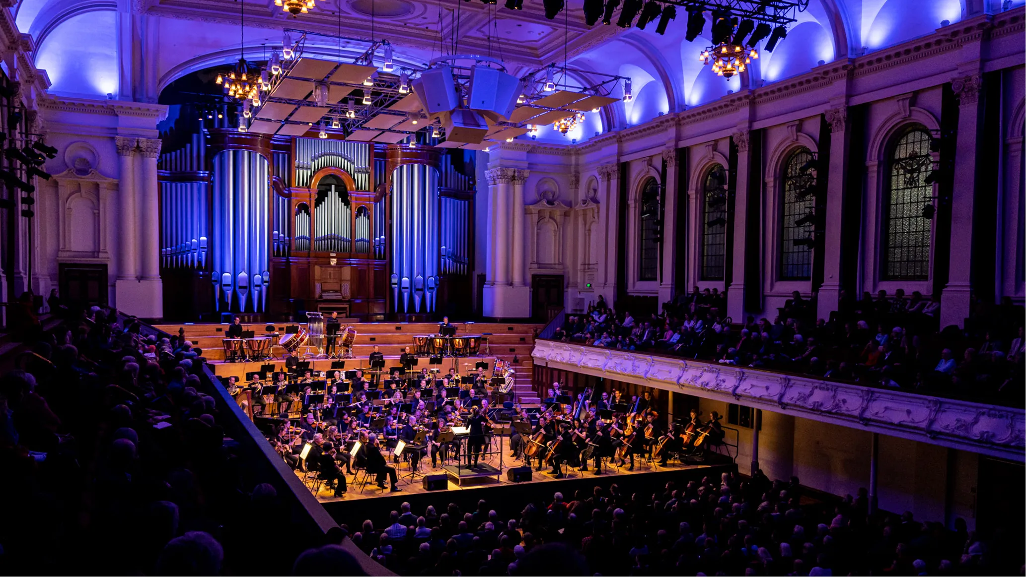 Orchestra performing in a grand hall with large organ pipes and audience seated in balcony and main floor.