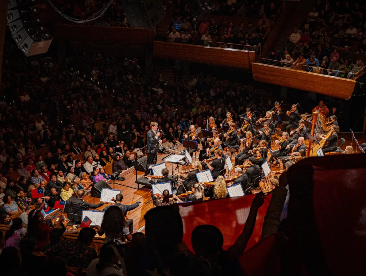 Orchestra performing on stage with conductor and audience watching in a concert hall.