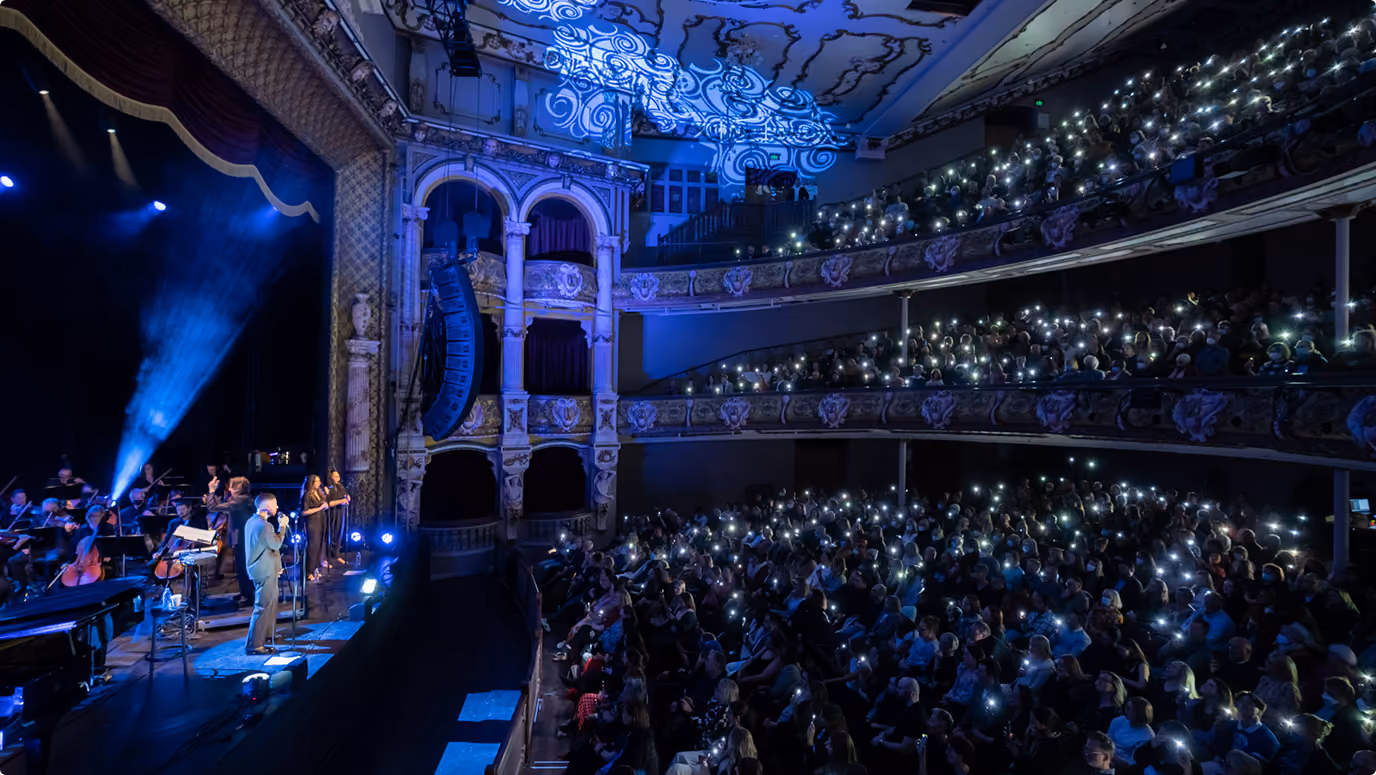 Crowded theater with a singer on stage and audience holding up lit phone lights.