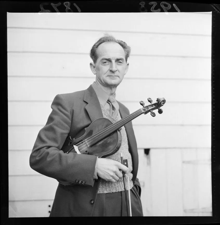 Black and white photo of a man in a suit holding a violin and bow standing against a wooden wall.