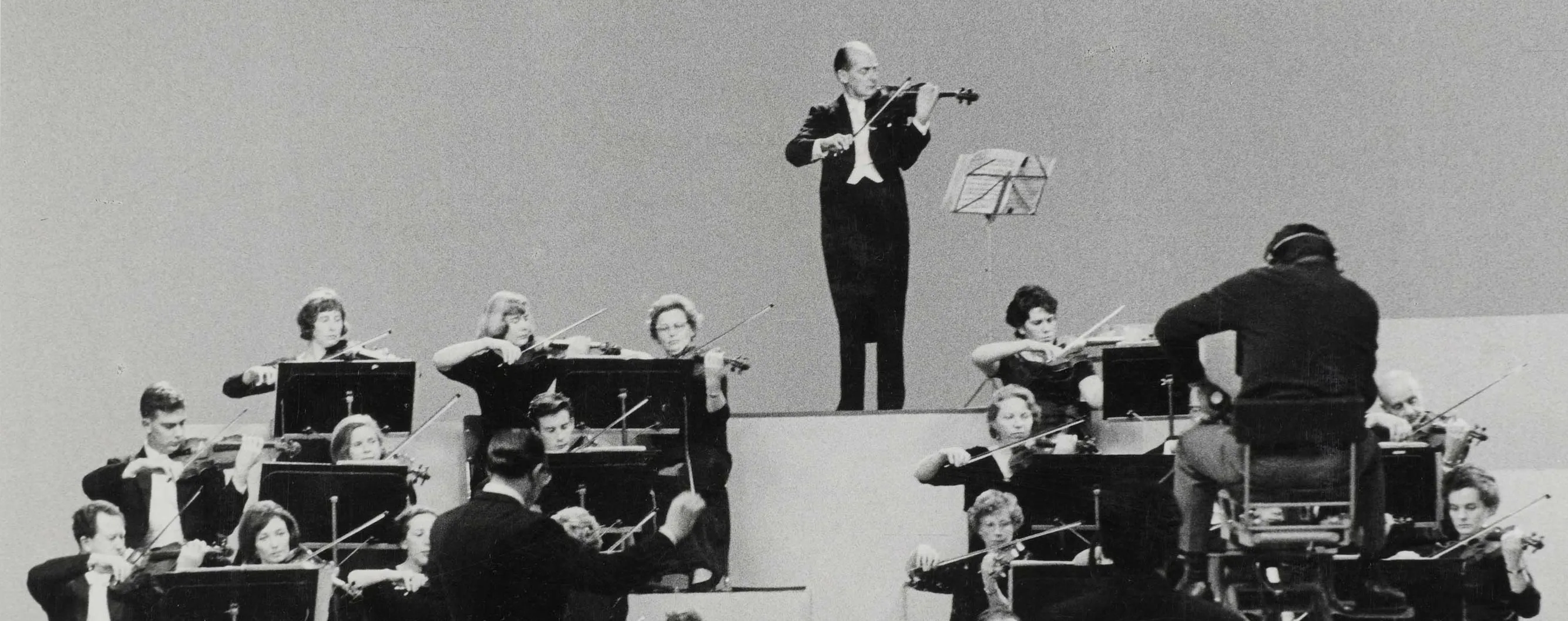 Black and white photo of violinists playing in an orchestra with a conductor and a solo violinist on a podium.