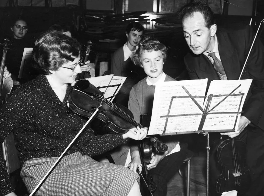 Black and white photo of a man instructing women playing violins while they read sheet music.