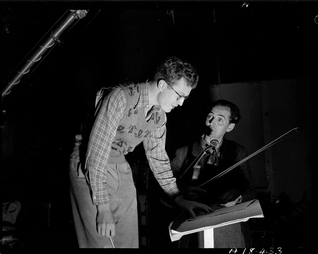 Two men in a dim-lit room with one holding a violin and the other pointing at sheet music.