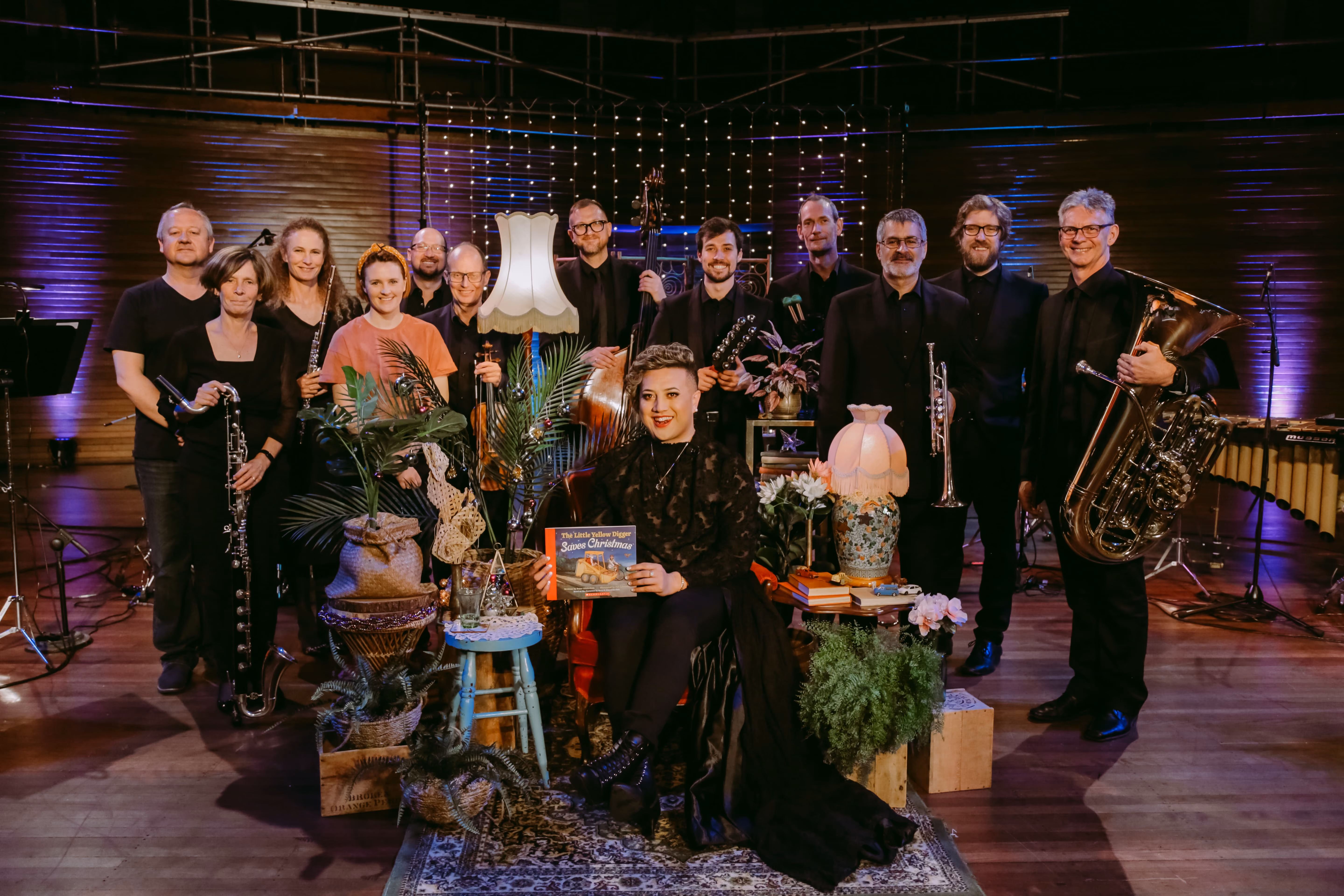 Group of musicians posing with instruments surrounded by plants, books, lamps, and a sitting woman holding a book.