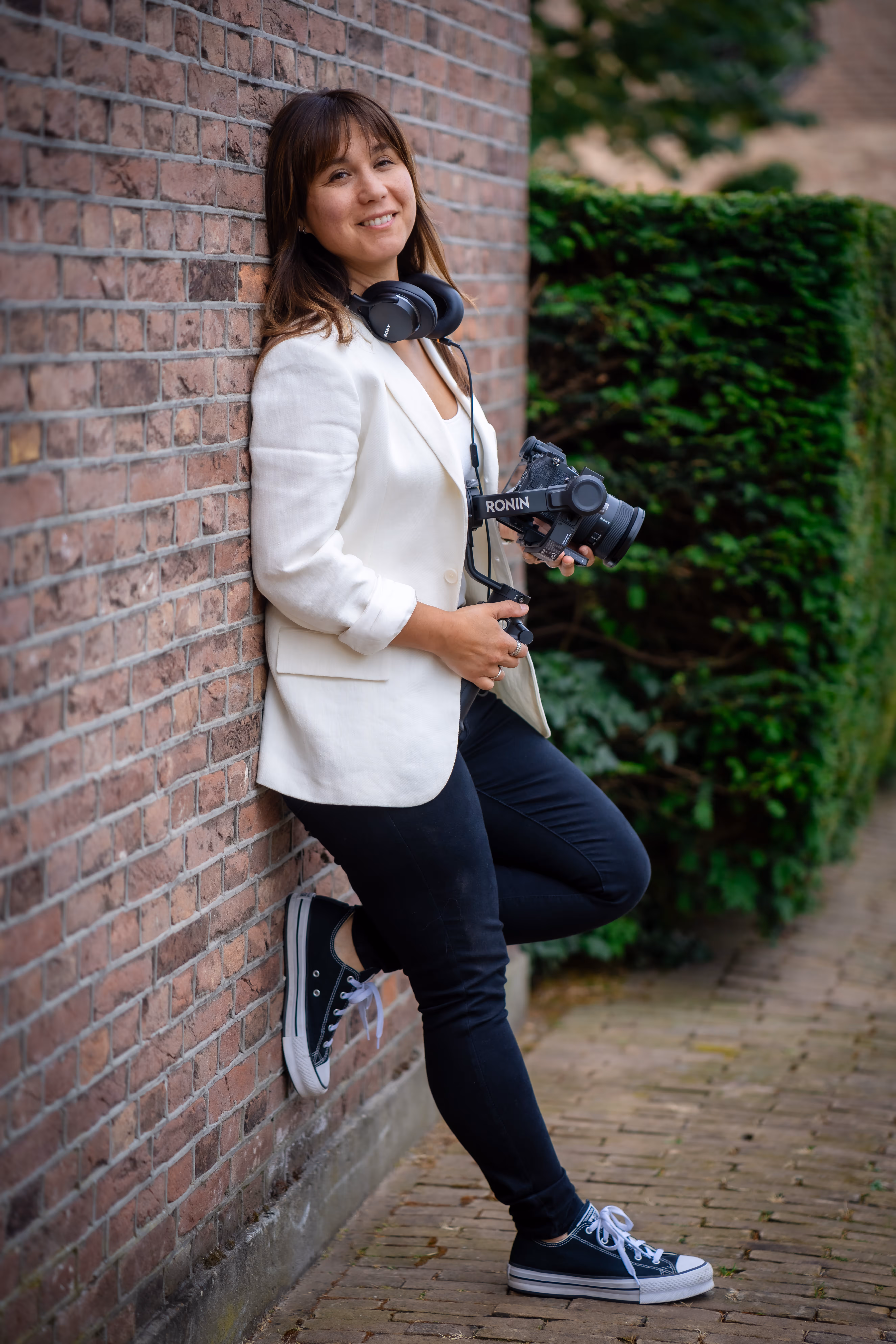 Lois Nieuwenhoven leunt tegen een bakstenen muur met haar camera en koptelefoon.