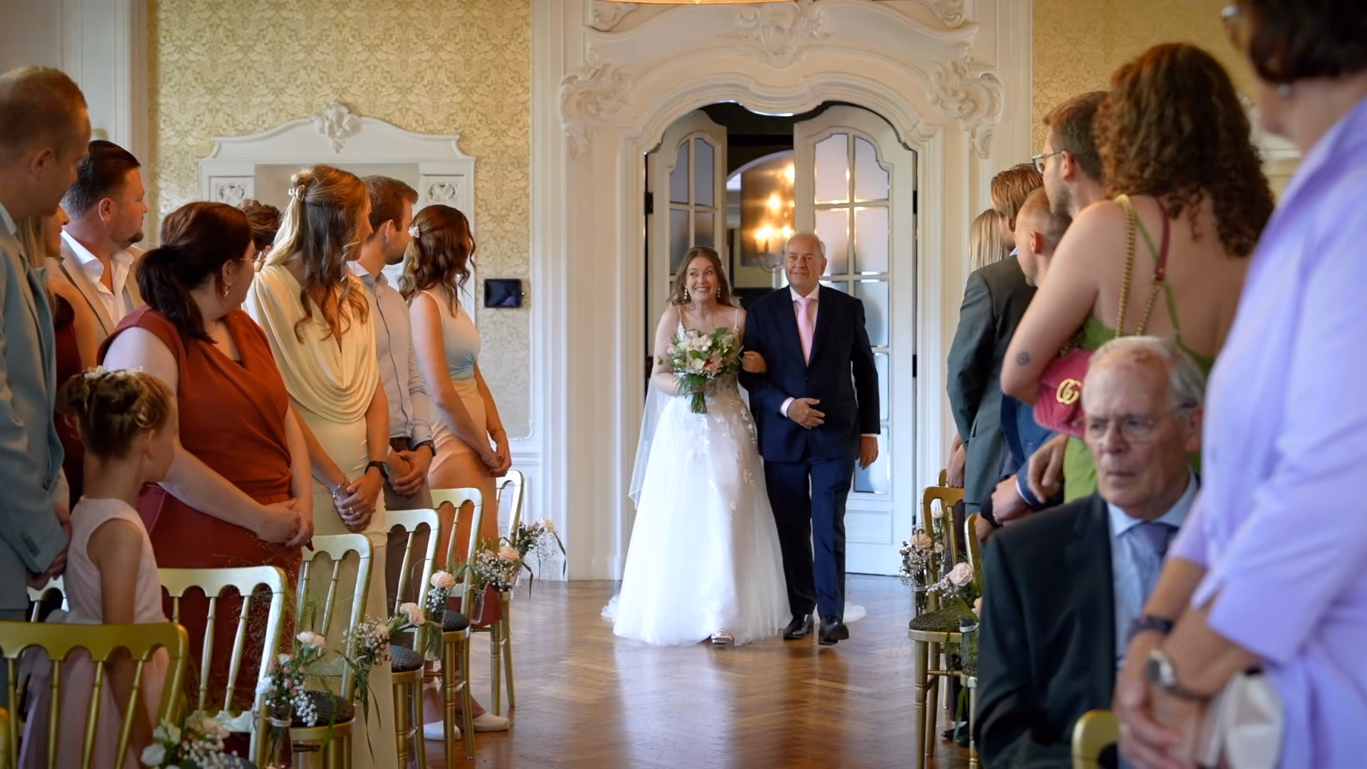 De bruid loopt aan de arm van een man tussen de gasten naar de ceremonie.