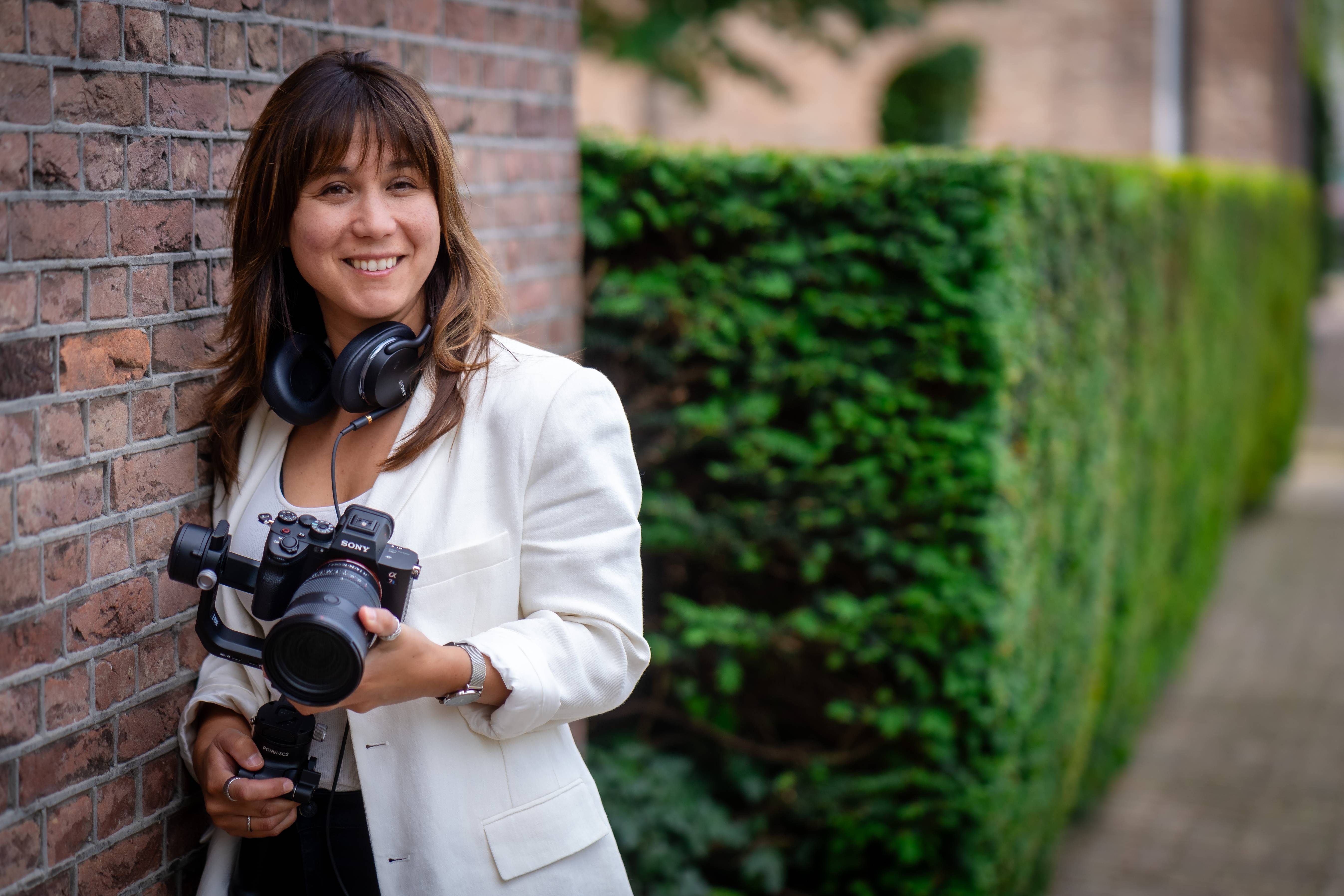 Lois Nieuwenhoven met haar camera en koptelefoon bij een bakstenen muur.