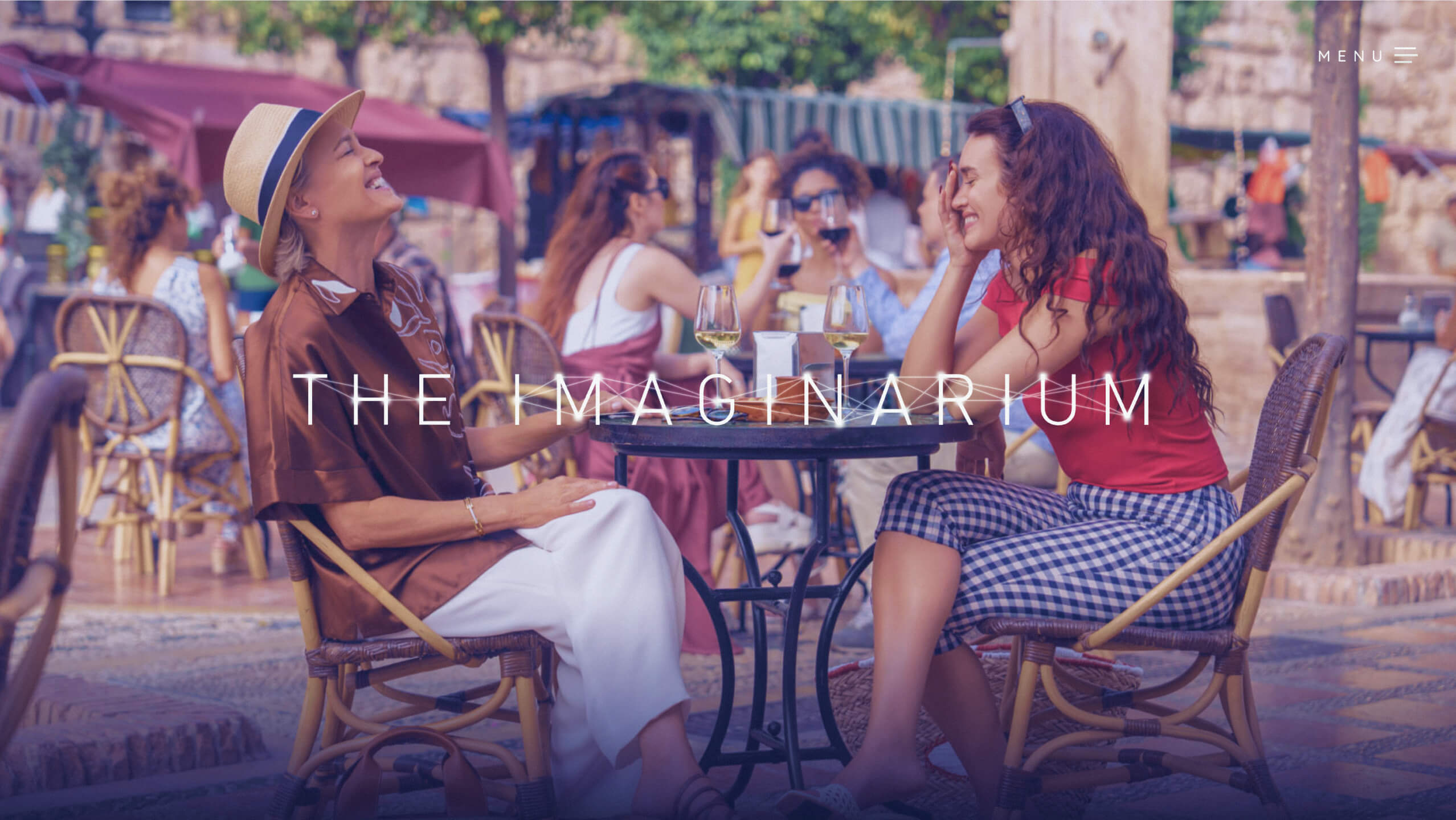 Two women sitting at an outdoor café table enjoying wine and laughing, surrounded by other seated people under umbrellas and trees in a sunny setting.